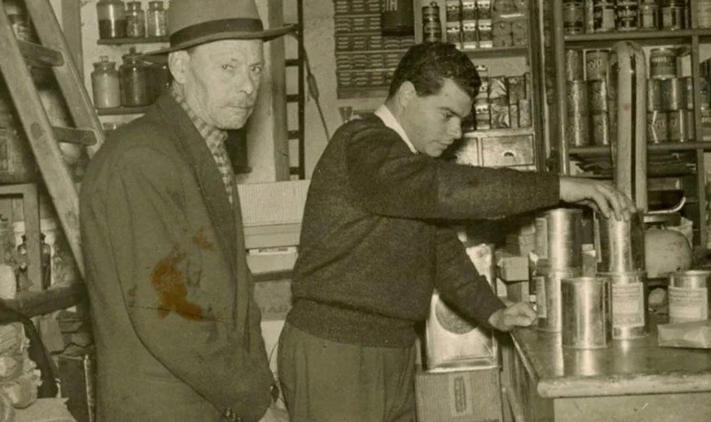 Due uomini in una cucina d'epoca, uno che mescola qualcosa in una pentola sui fornelli mentre l'altro lo osserva.