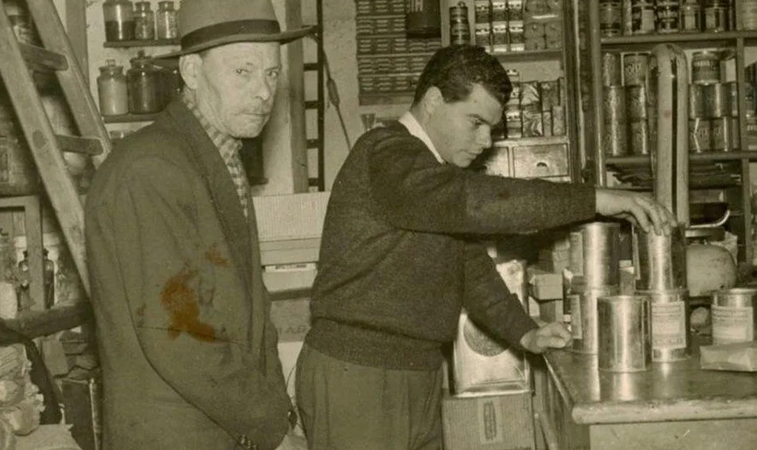 Due uomini in una cucina d'epoca, uno che mescola qualcosa in una pentola sui fornelli mentre l'altro lo osserva.