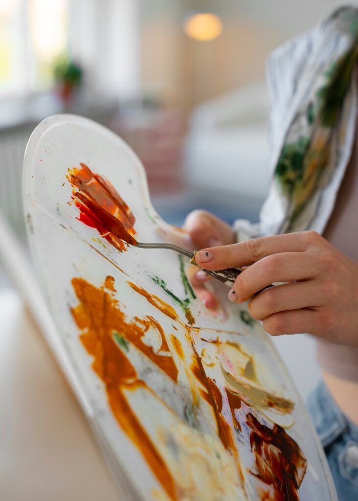 Mani che dipingono con colori arancione, rosso e marrone su una tela bianca in uno studio luminoso.