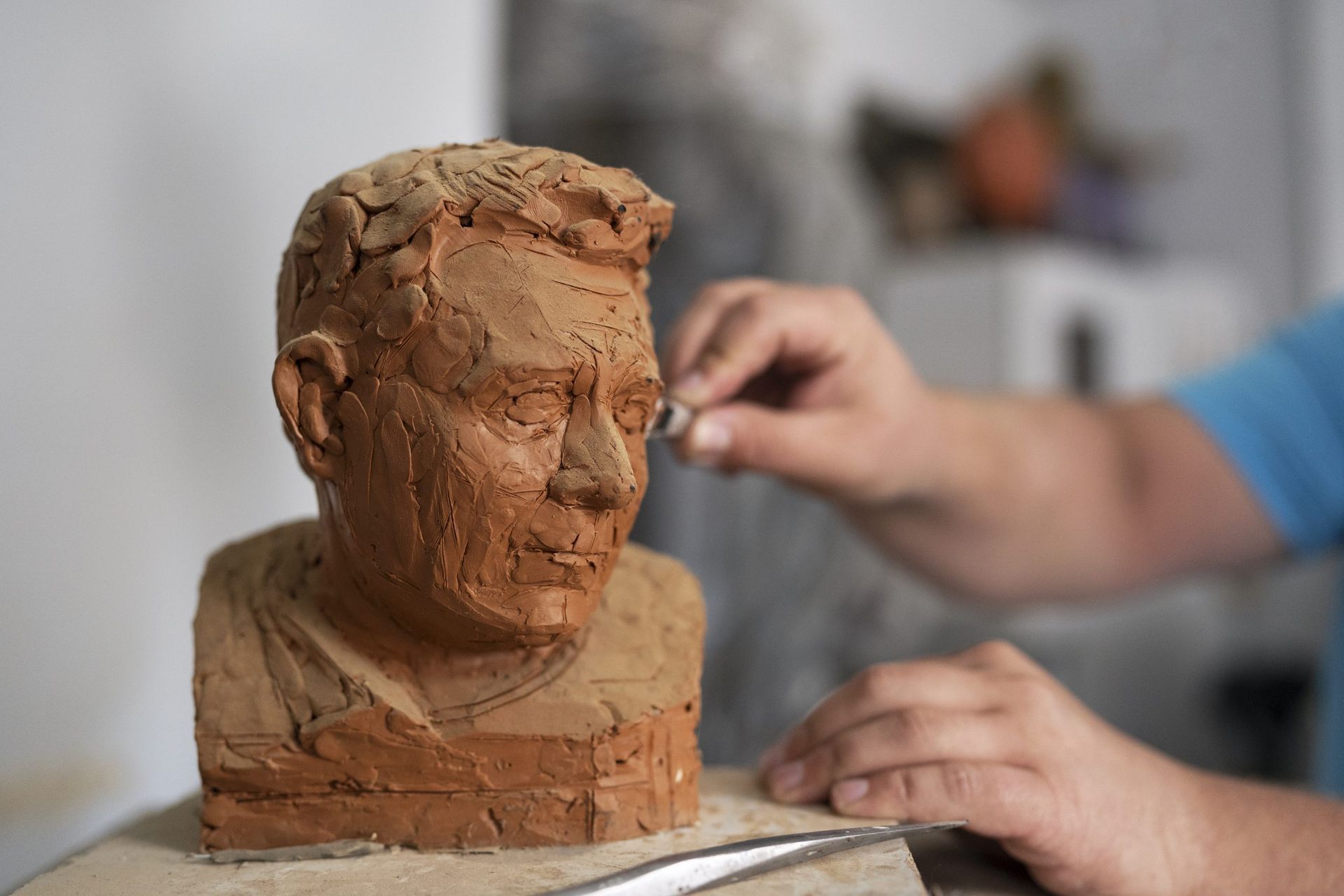 Mani che modellano un busto di argilla su un banco da lavoro, con un artista sfocato sullo sfondo.