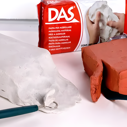 Materiali artistici con una scatola di argilla DAS rossa, argilla bianca e strumenti con manico blu su una superficie bianca.