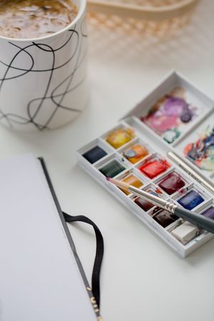 Un set di acquerelli, un pennello, un album da disegno e una tazza da caffè su un tavolo bianco.