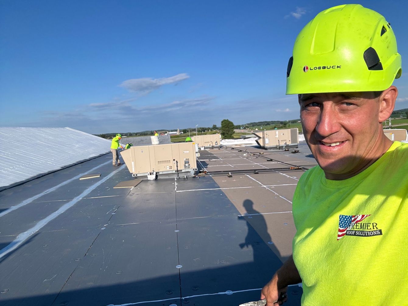 Man in hardhat and yellow shirt smiles on a roof; another person works in the background.