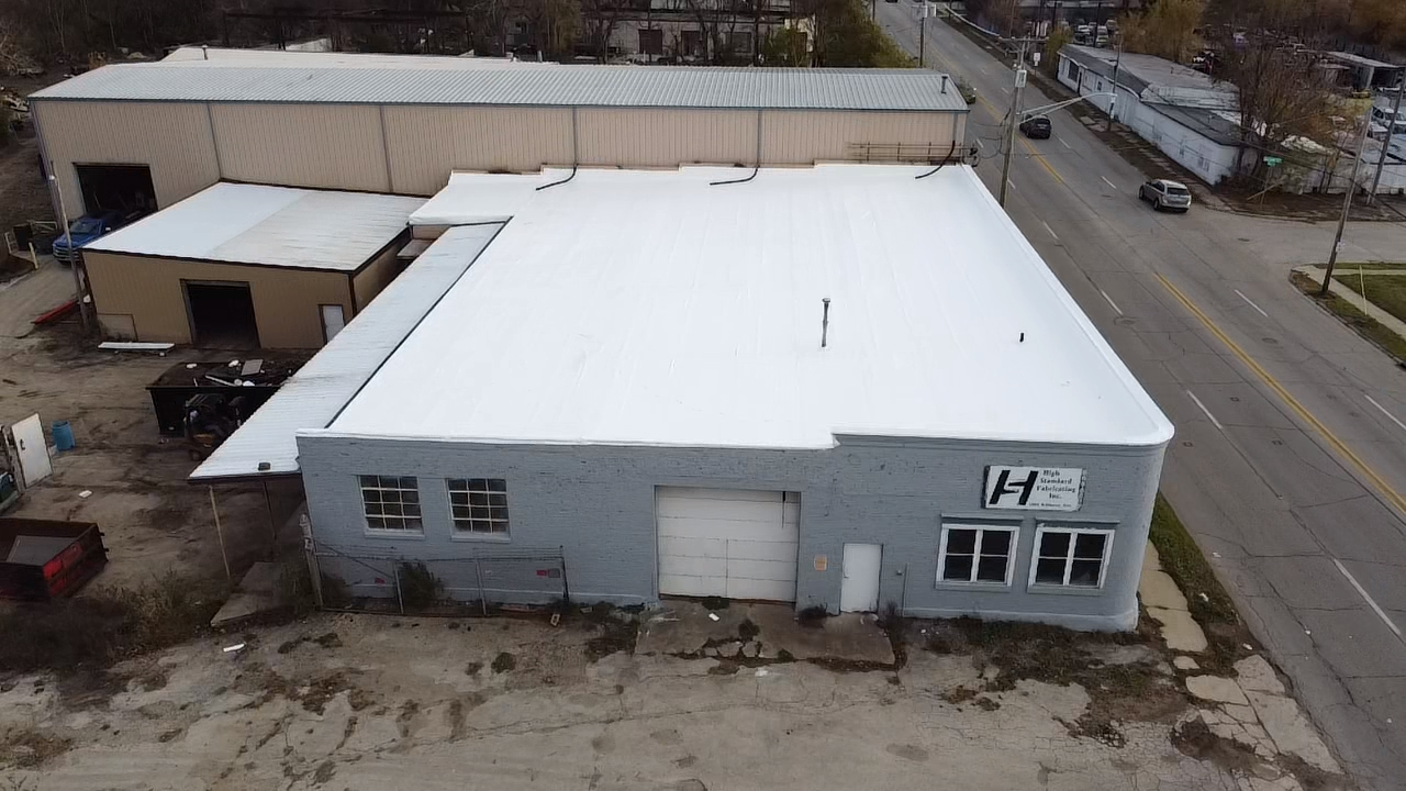 Aerial view of a gray building with a white roof, next to a street with cars and other buildings.