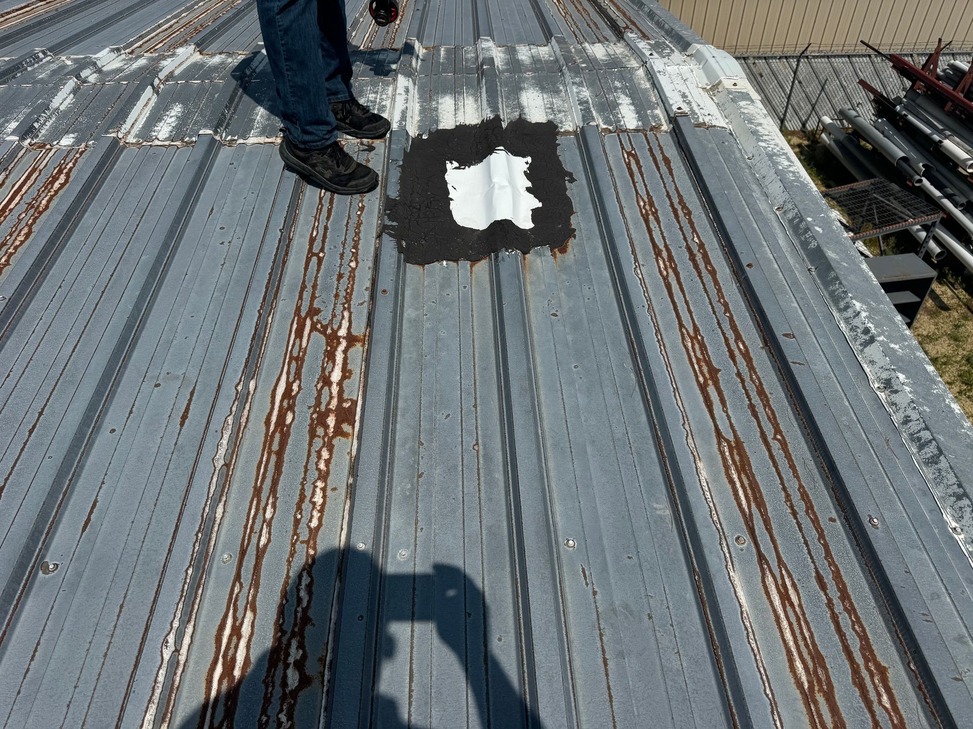Person on weathered metal roof, applying sealant to a patch. Grey and rust colors.