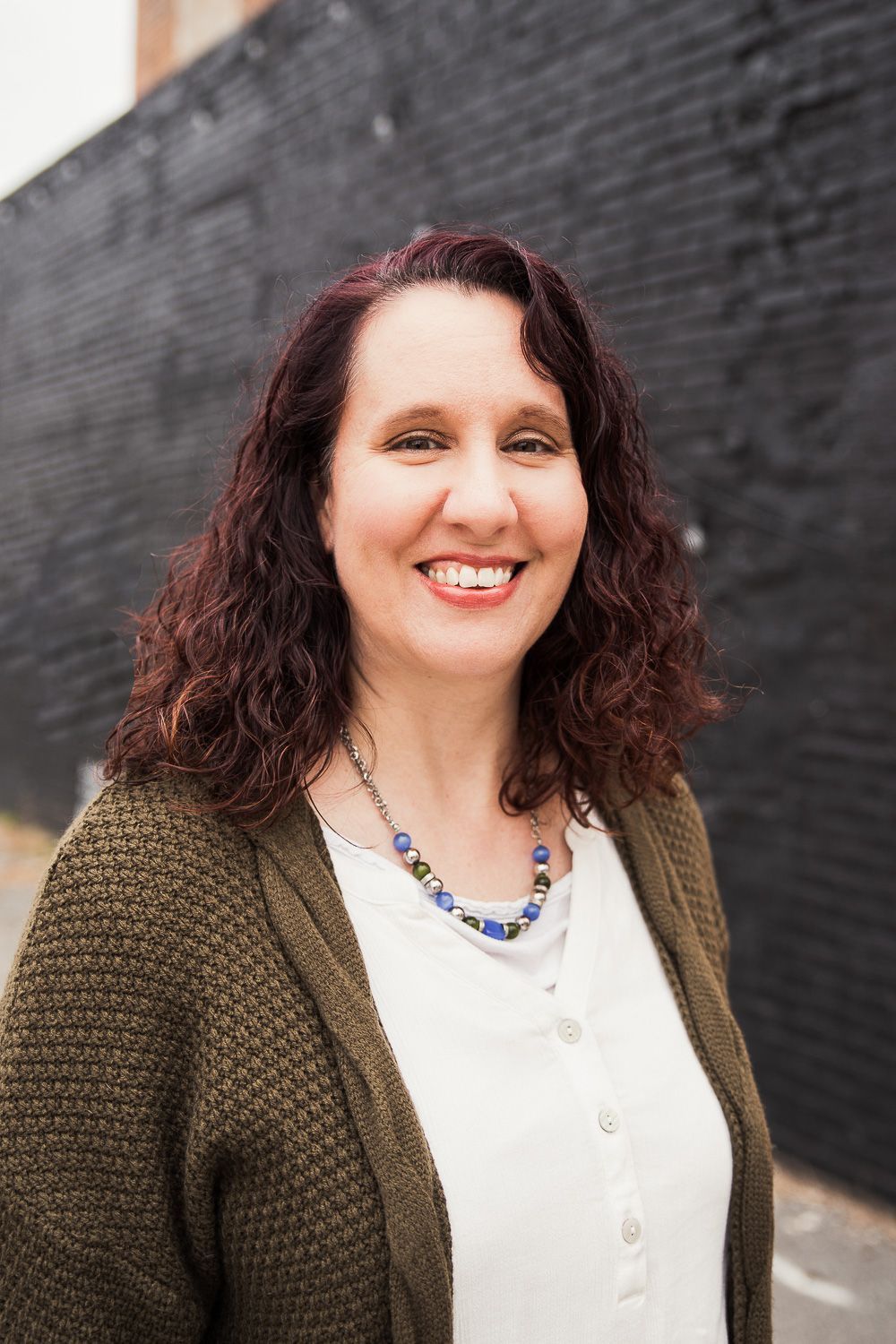 A woman wearing a green cardigan and a blue necklace is smiling in front of a black wall.