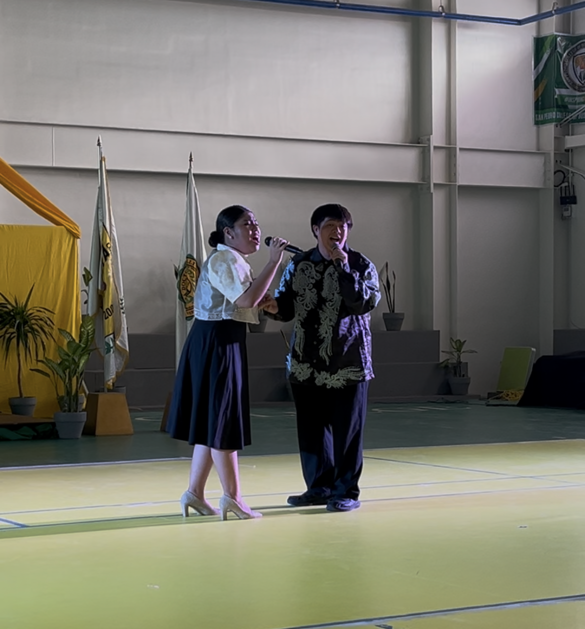 Two people speaking on a stage, one holding a microphone in a gymnasium-like hall.
