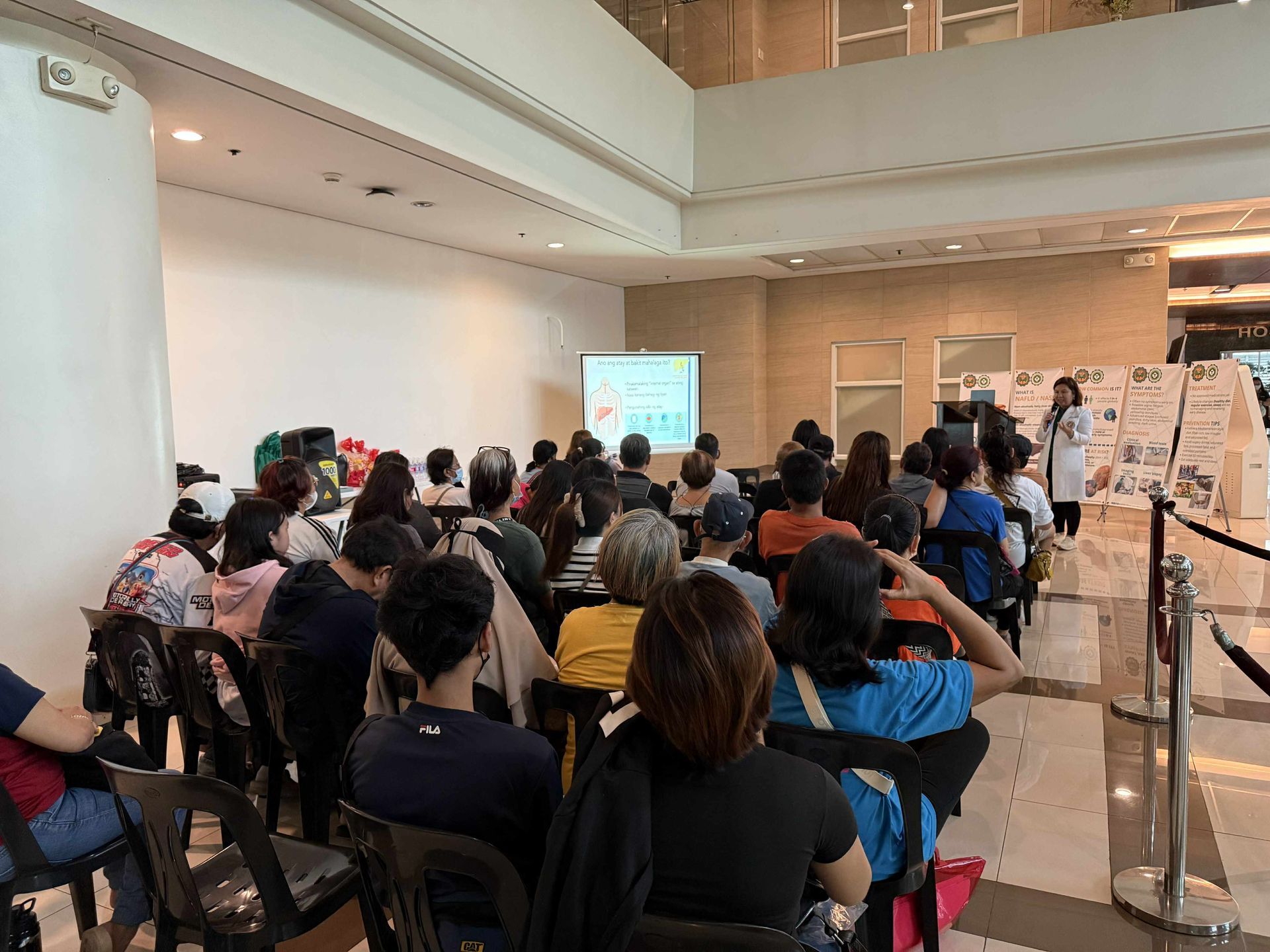 Audience seated in a white conference hall watching a presentation on a screen at the front.