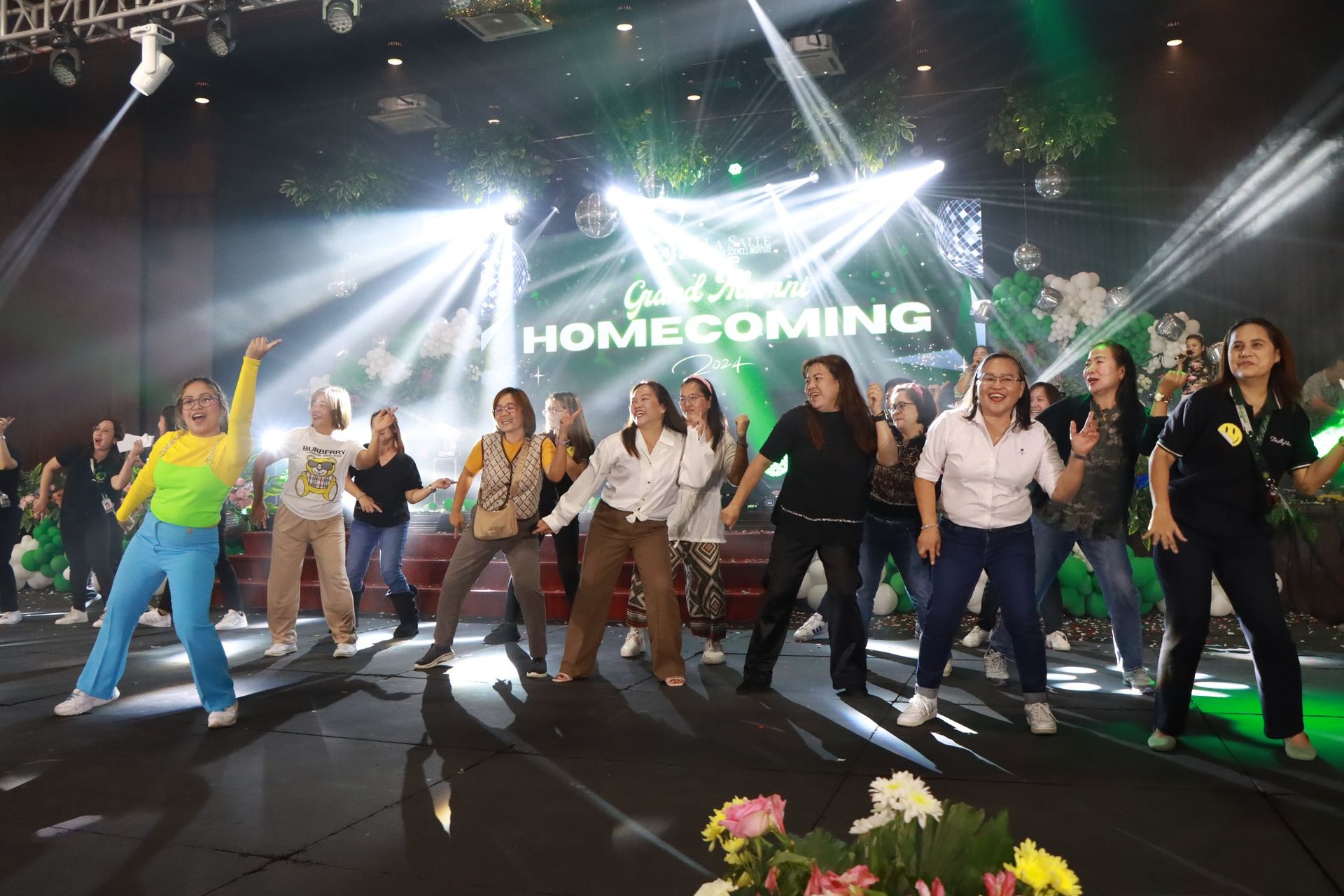 Group dancing on stage under bright lights, with “HOMECOMING” displayed on the screen behind them.