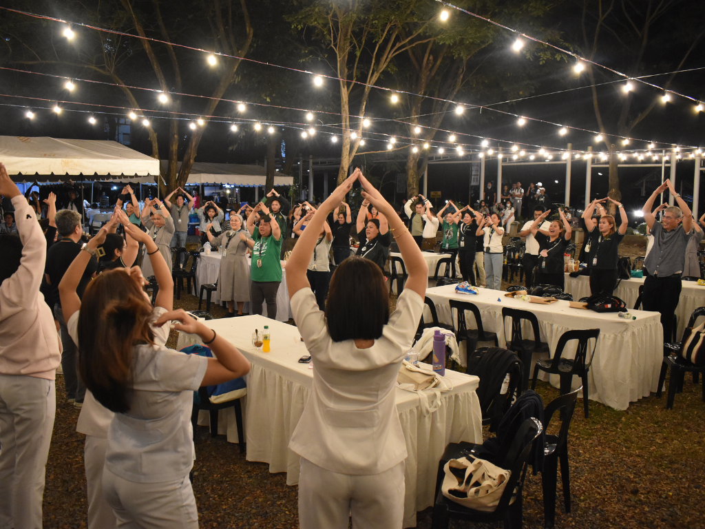 Outdoor evening gathering with people raising their arms under string lights at banquet tables