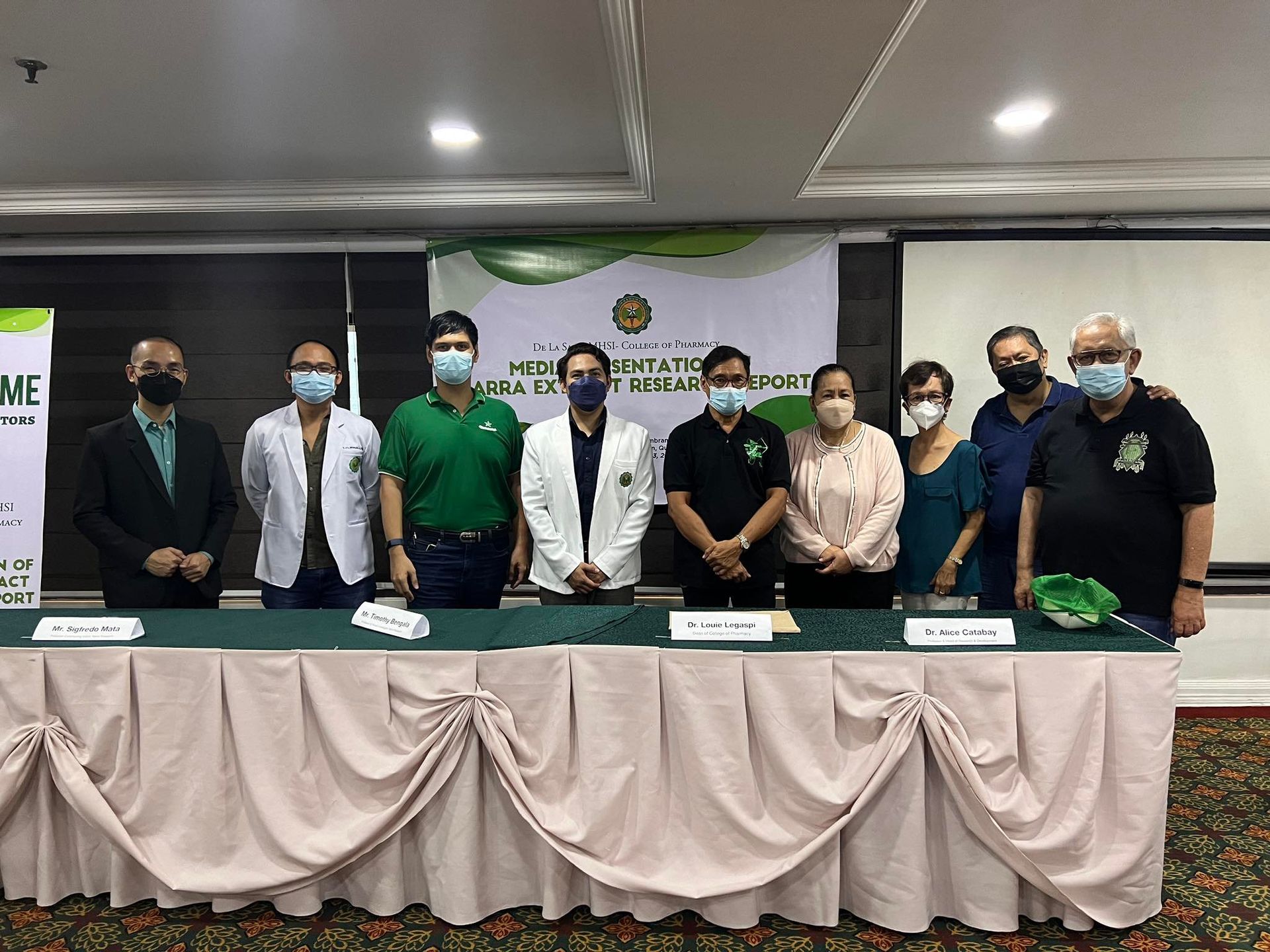 Nine masked people standing behind a panel table at a formal indoor event