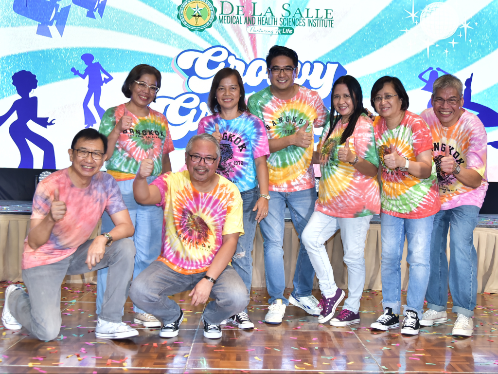 Group of smiling people in tie-dye shirts posing on a stage with a colorful mural backdrop