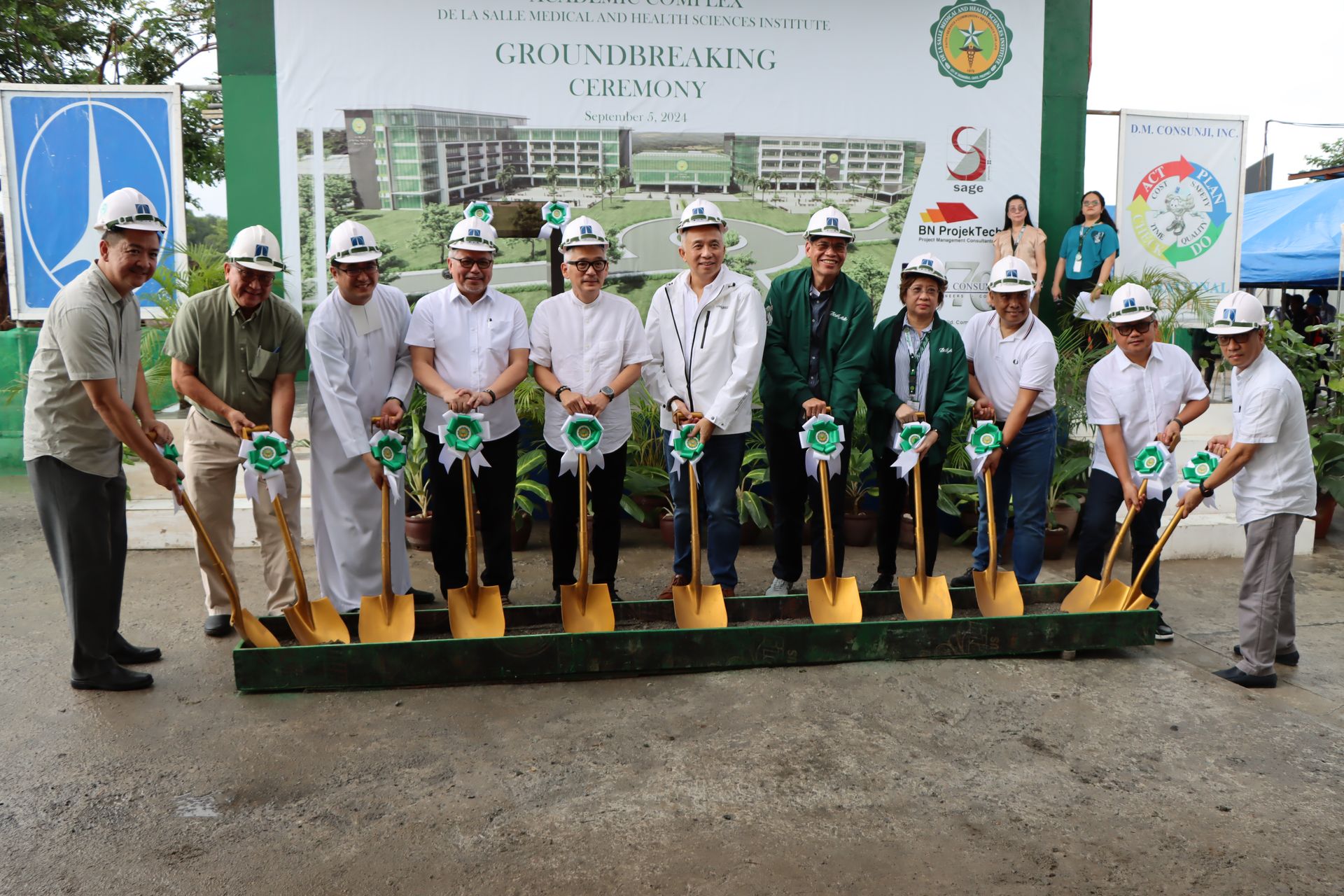 Officials in white uniforms hold shovels during a groundbreaking ceremony before a bannered building site.