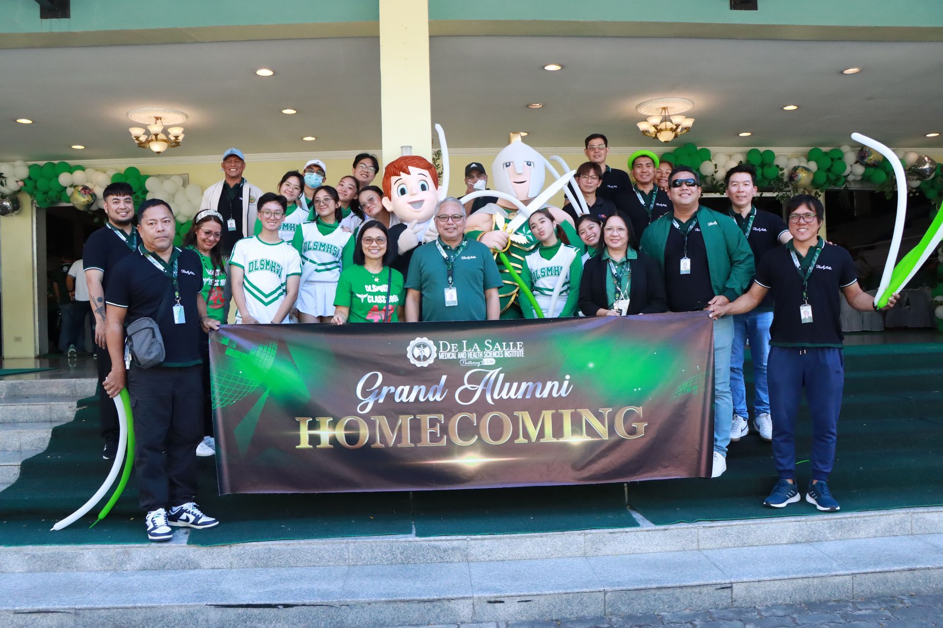 Group of people in green shirts posing with a “Grand Alumni Homecoming” banner outside a building