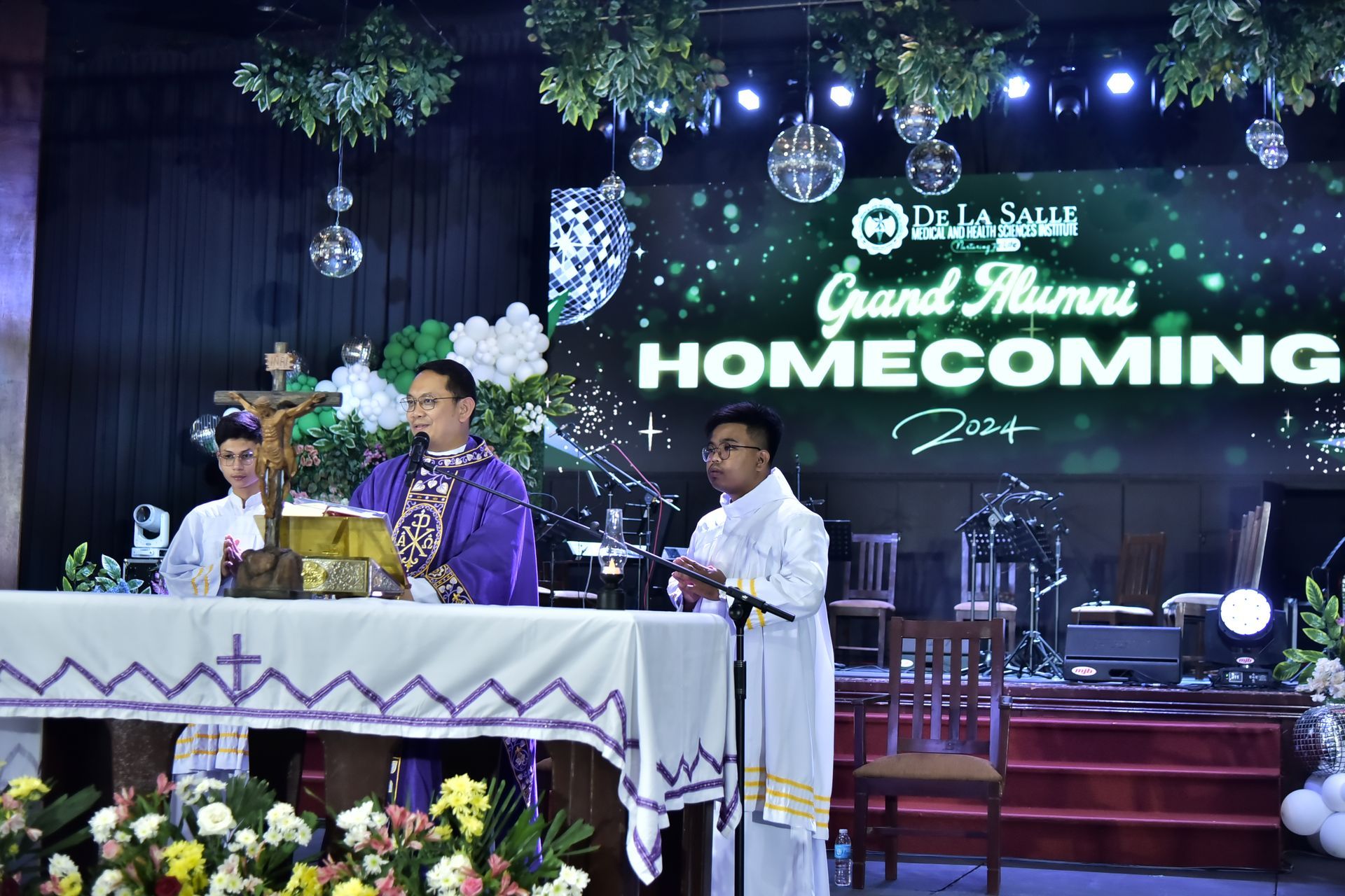 Three men at a decorated stage podium during a “Grand Family Homecoming” event with floral arrangements and lights