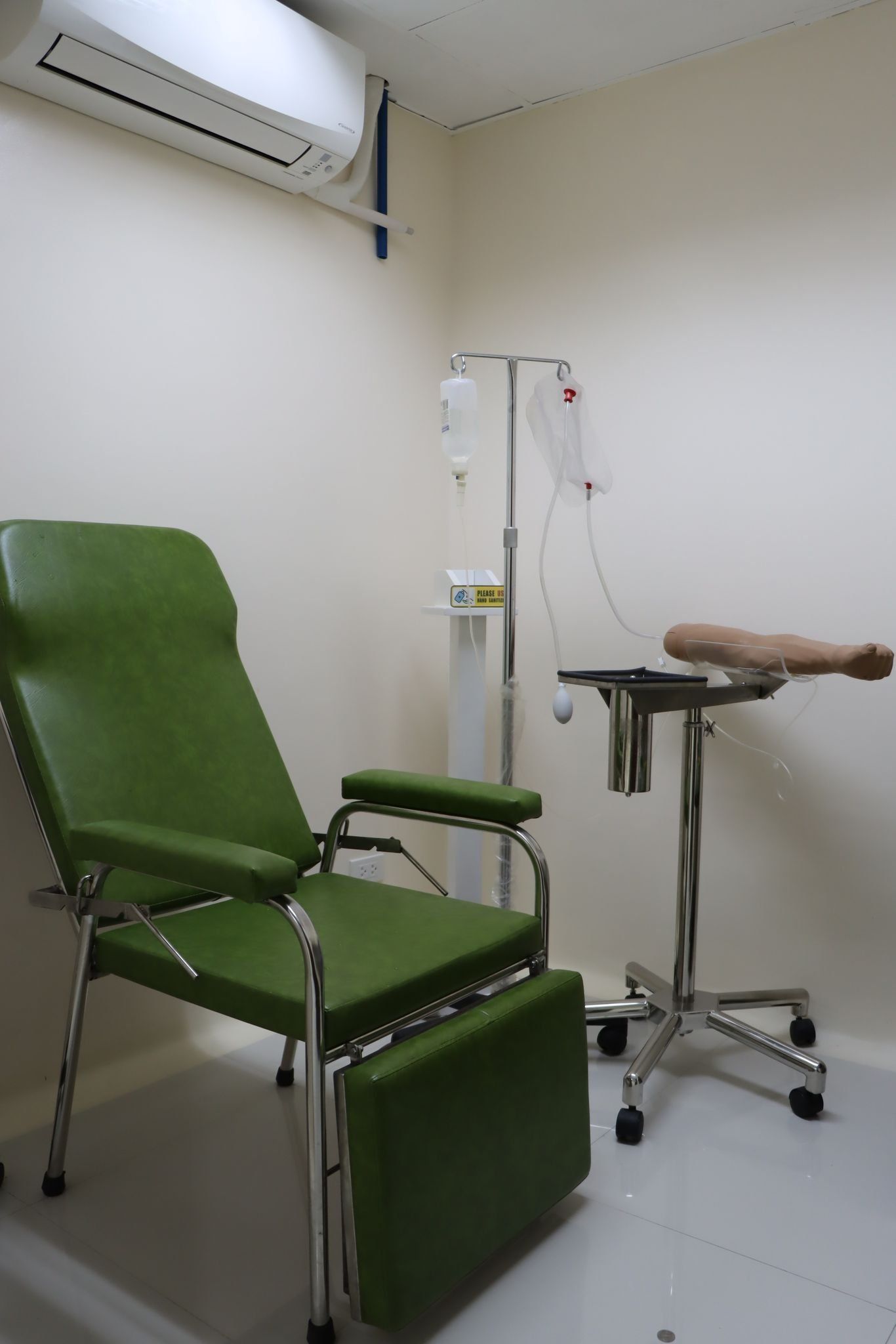 Green medical examination chair and stool in a small clinic room with white walls.