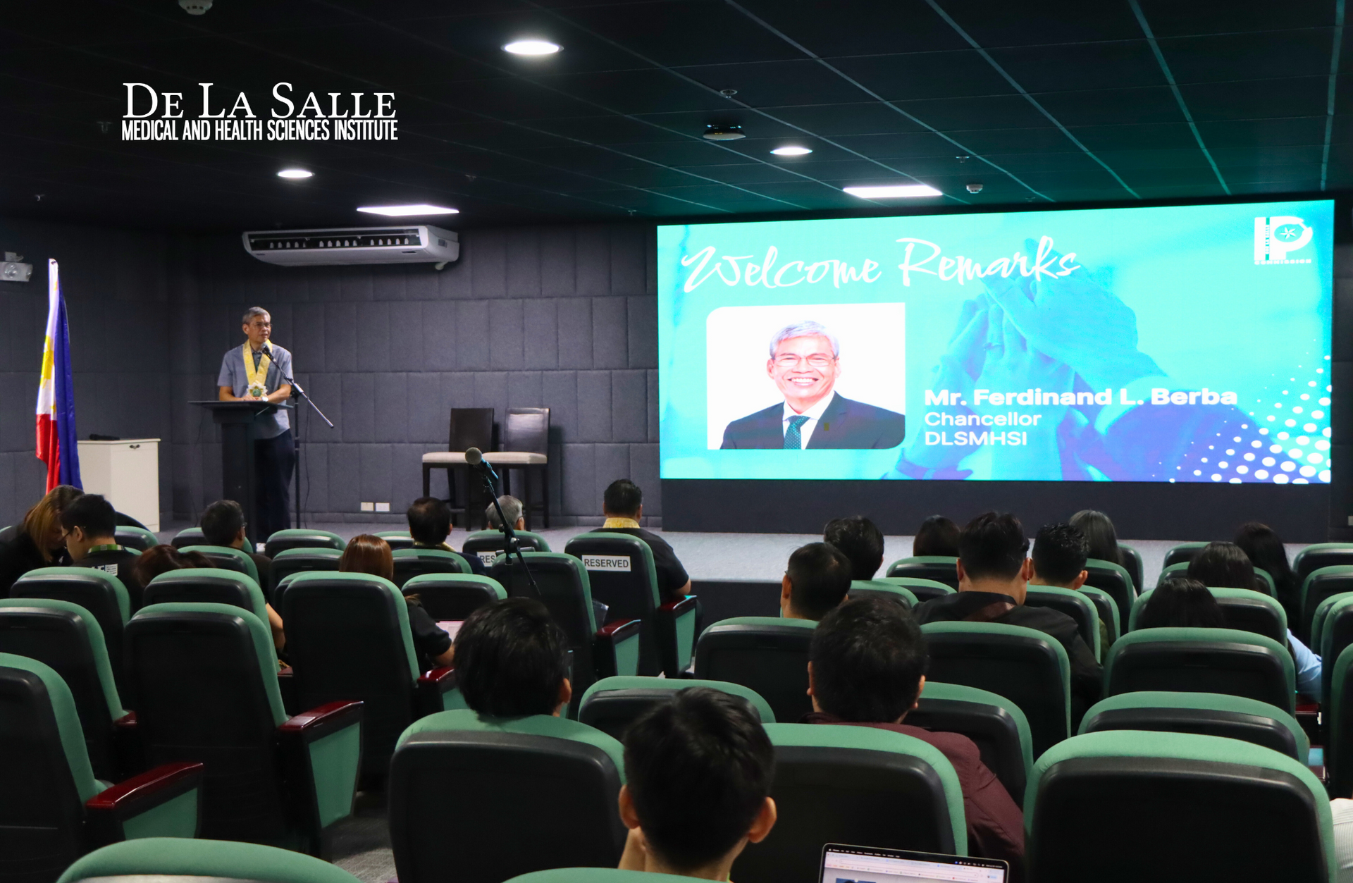 Lecture hall with a speaker at podium and a large presentation screen in a mostly empty auditorium