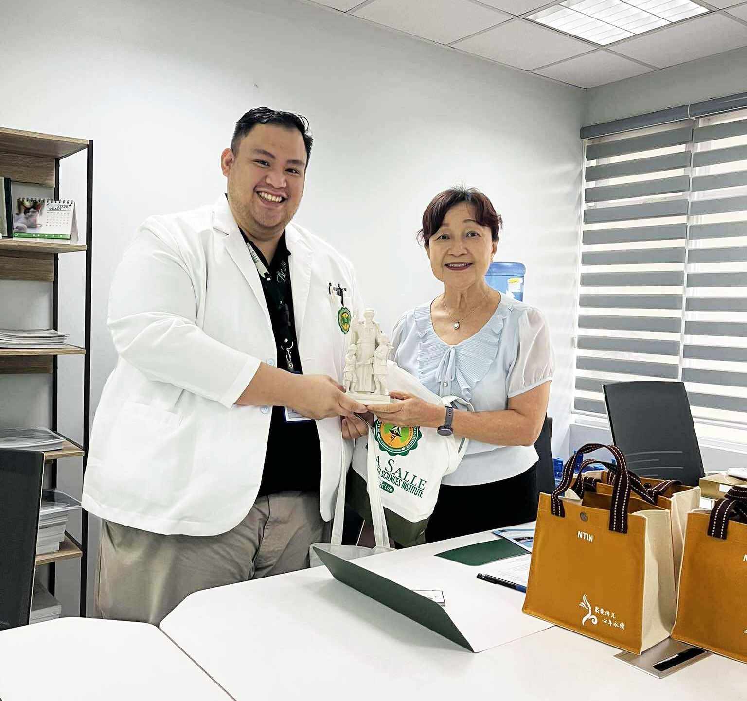 Two people exchanging a gift in a bright office, with bags on a desk and smiling expressions
