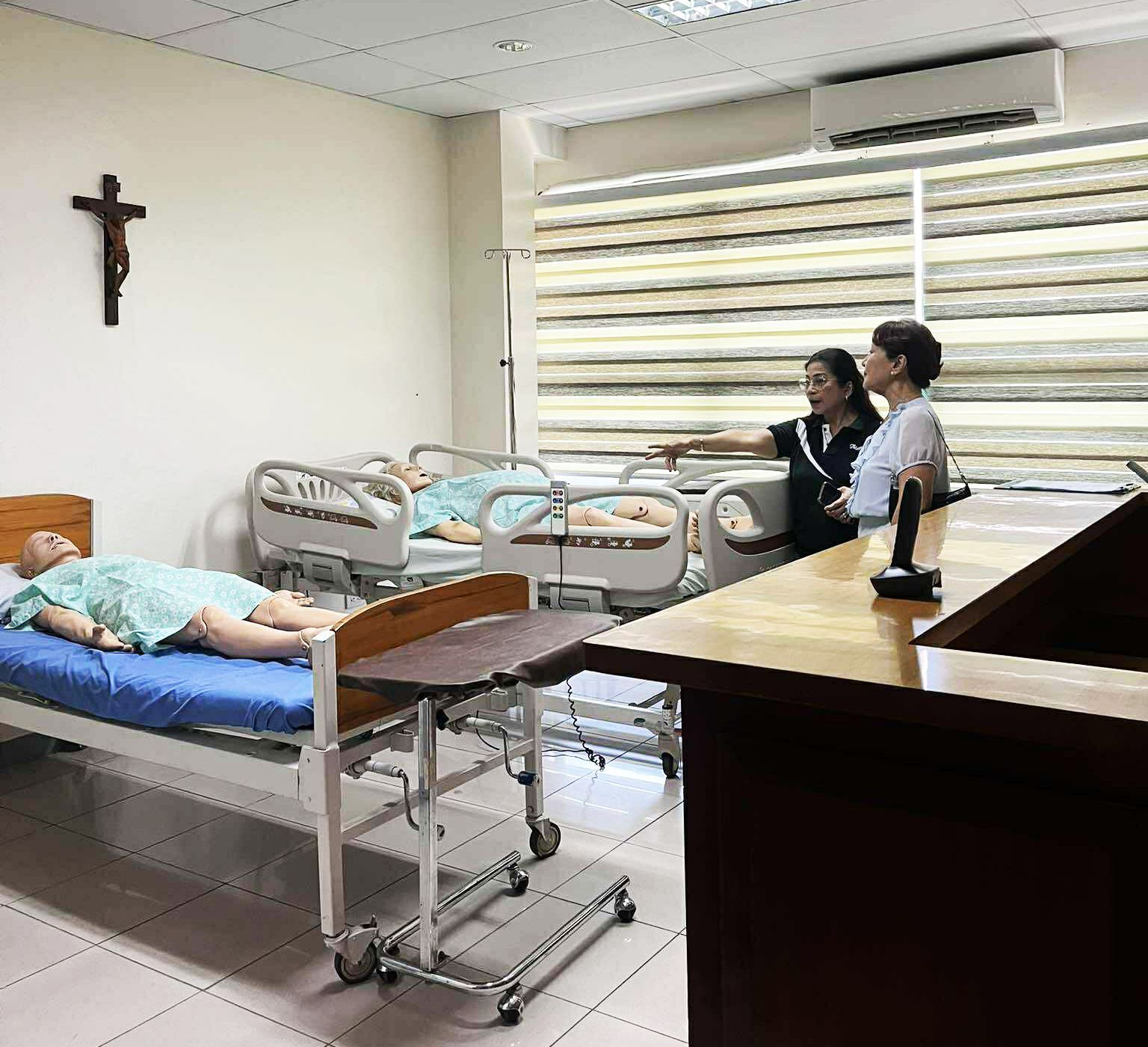 Hospital room with a patient in bed, two people standing nearby, and a crucifix on the wall.