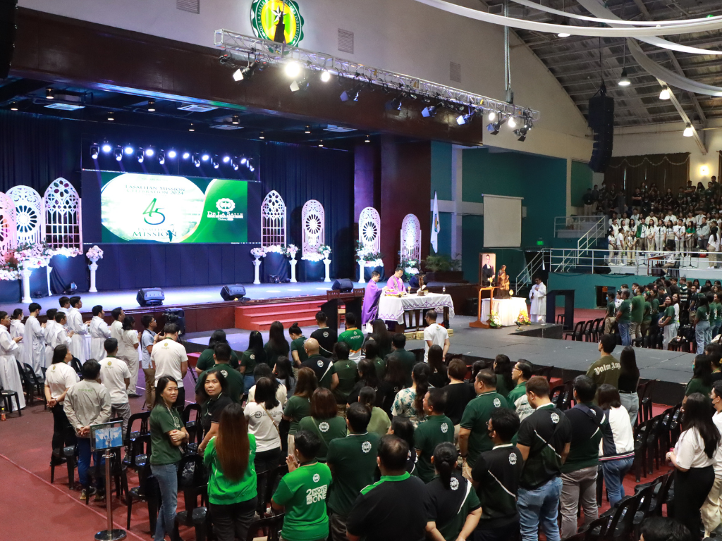 Large indoor event with a stage, green screen backdrop, and a seated audience in matching shirts