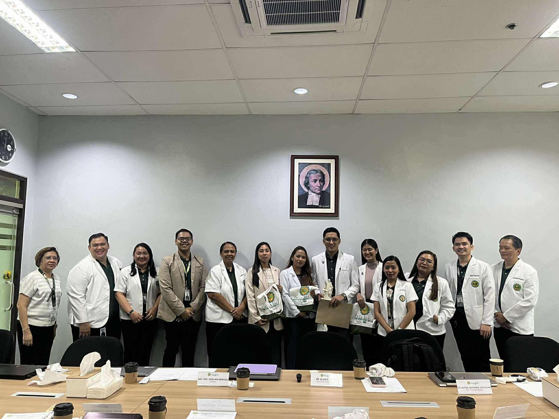 Group of people in white coats posing in a conference room with a framed portrait on the wall.
