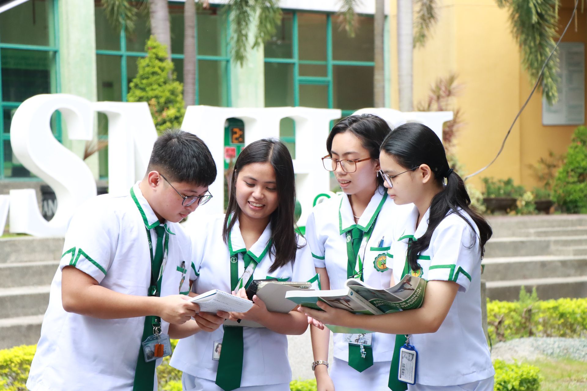 Four students in white-green uniforms study together outdoors on a campus lawn.