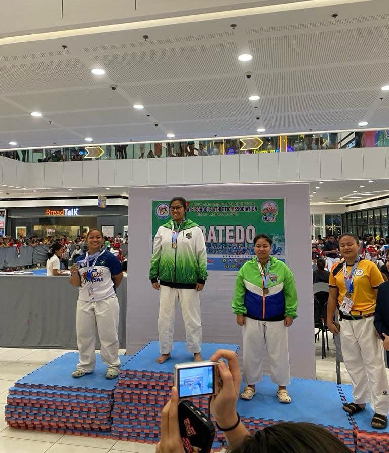 Podium ceremony with four medalists on a blue stage in a mall, standing before a “CHAMPIONS” backdrop.