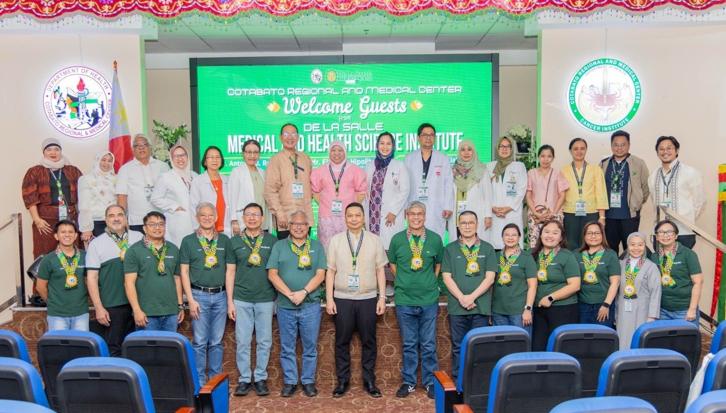 Group of people posing on stage in front of a green event banner, some seated in front and others standing behind.