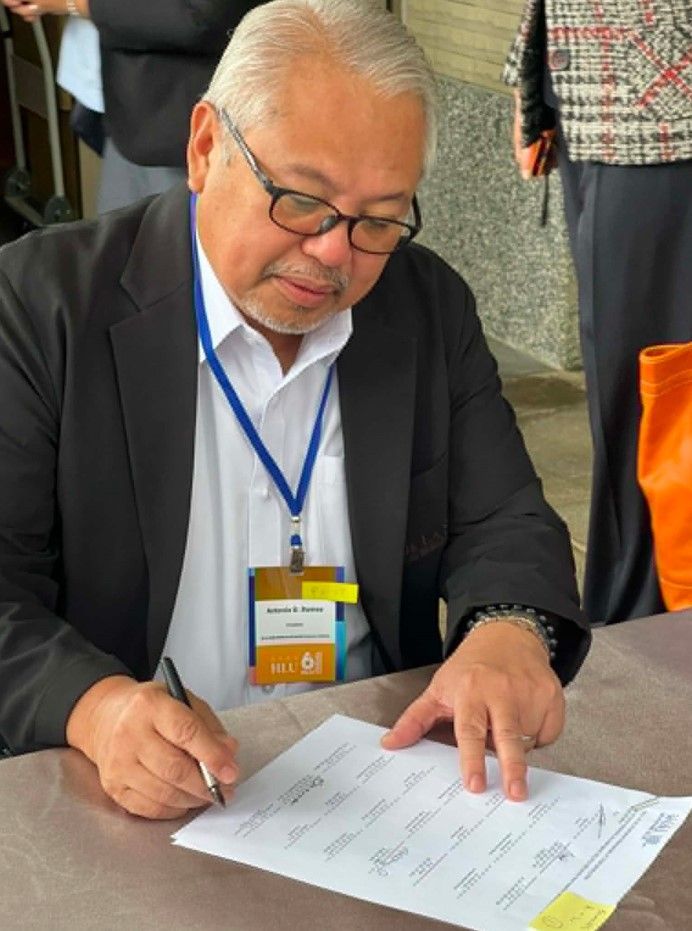Man in black blazer signing a document at a table, wearing glasses and a blue lanyard
