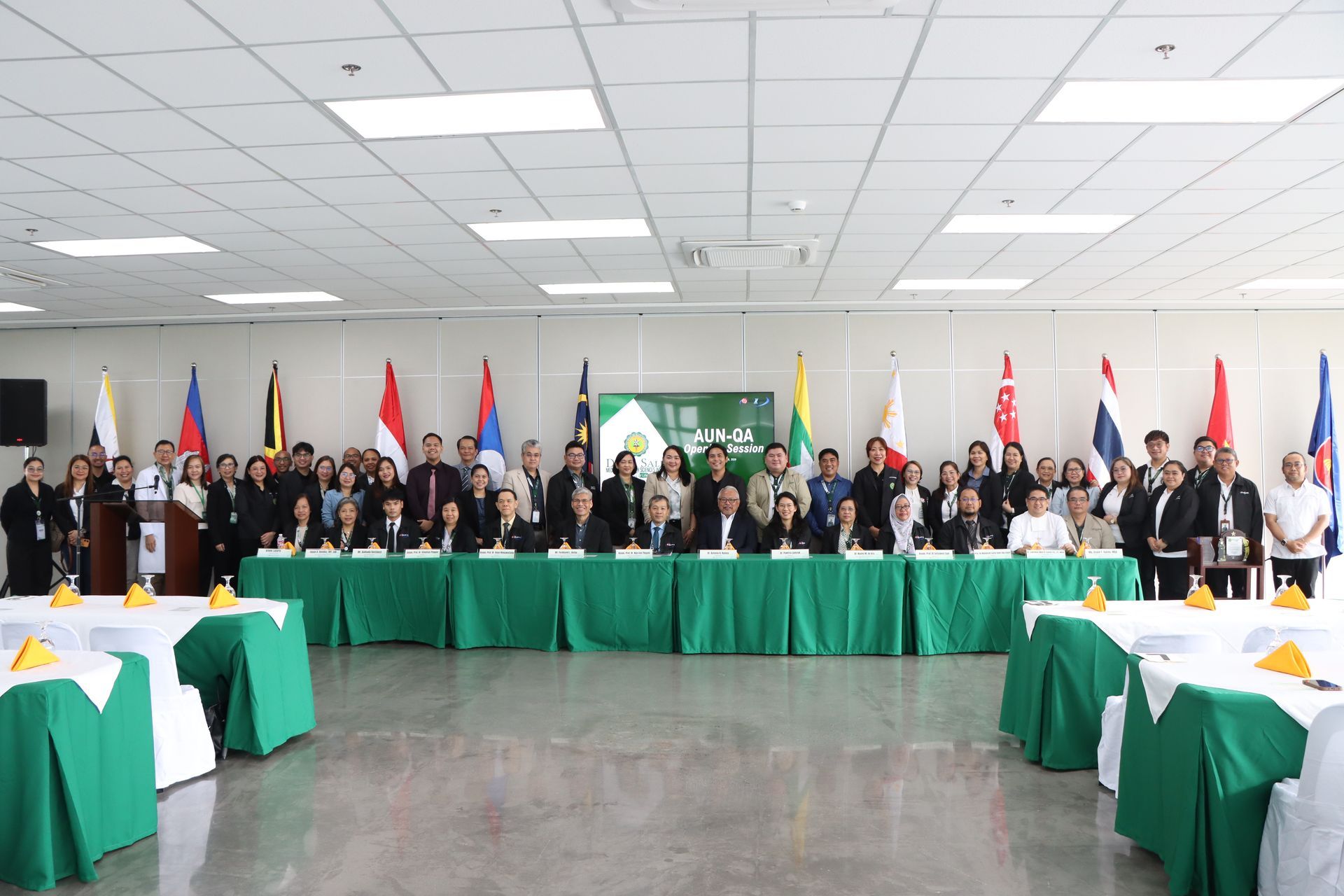 Group of people posing behind green-covered tables in a conference room with national flags in the background