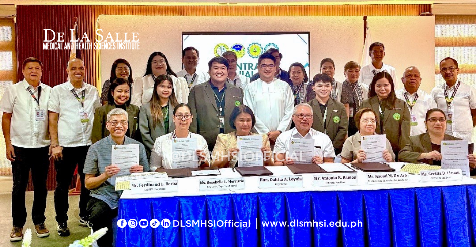 Group of people posing at a formal signing ceremony behind a blue table with banners and a white backdrop