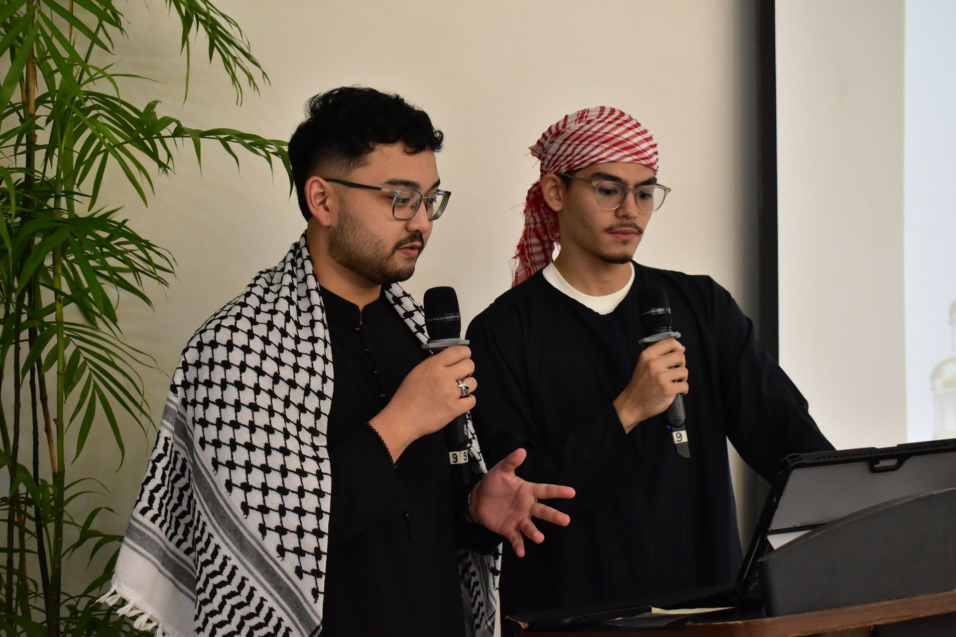 Two speakers holding microphones stand at a podium, one in a black shirt and one in a black robe with a red-and-white headscarf.
