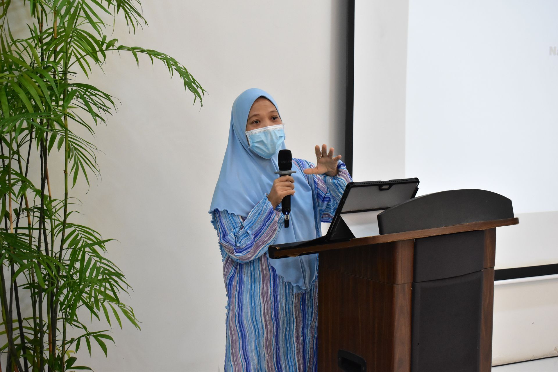 Woman in a blue hijab speaking at a podium with a microphone, beside an open laptop.