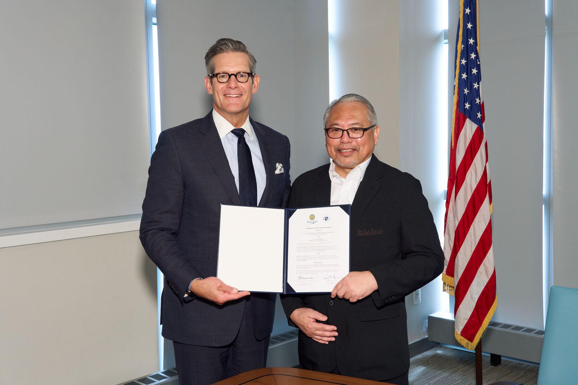 Two men in suits hold documents at a formal office ceremony, with a U.S. flag in the background