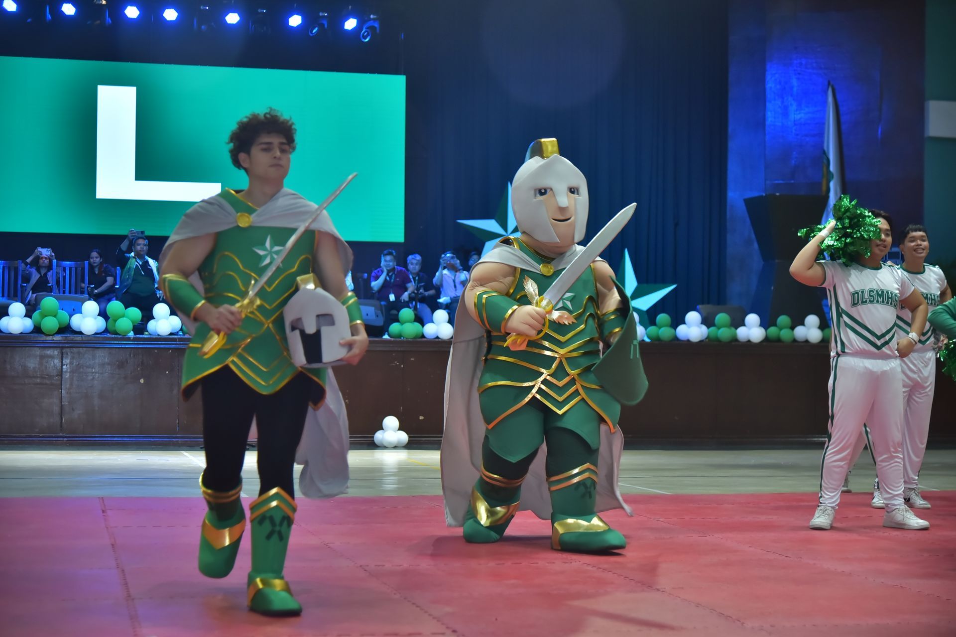 Three costumed performers on a stage, including two in green outfits holding props, with a large screen behind them.