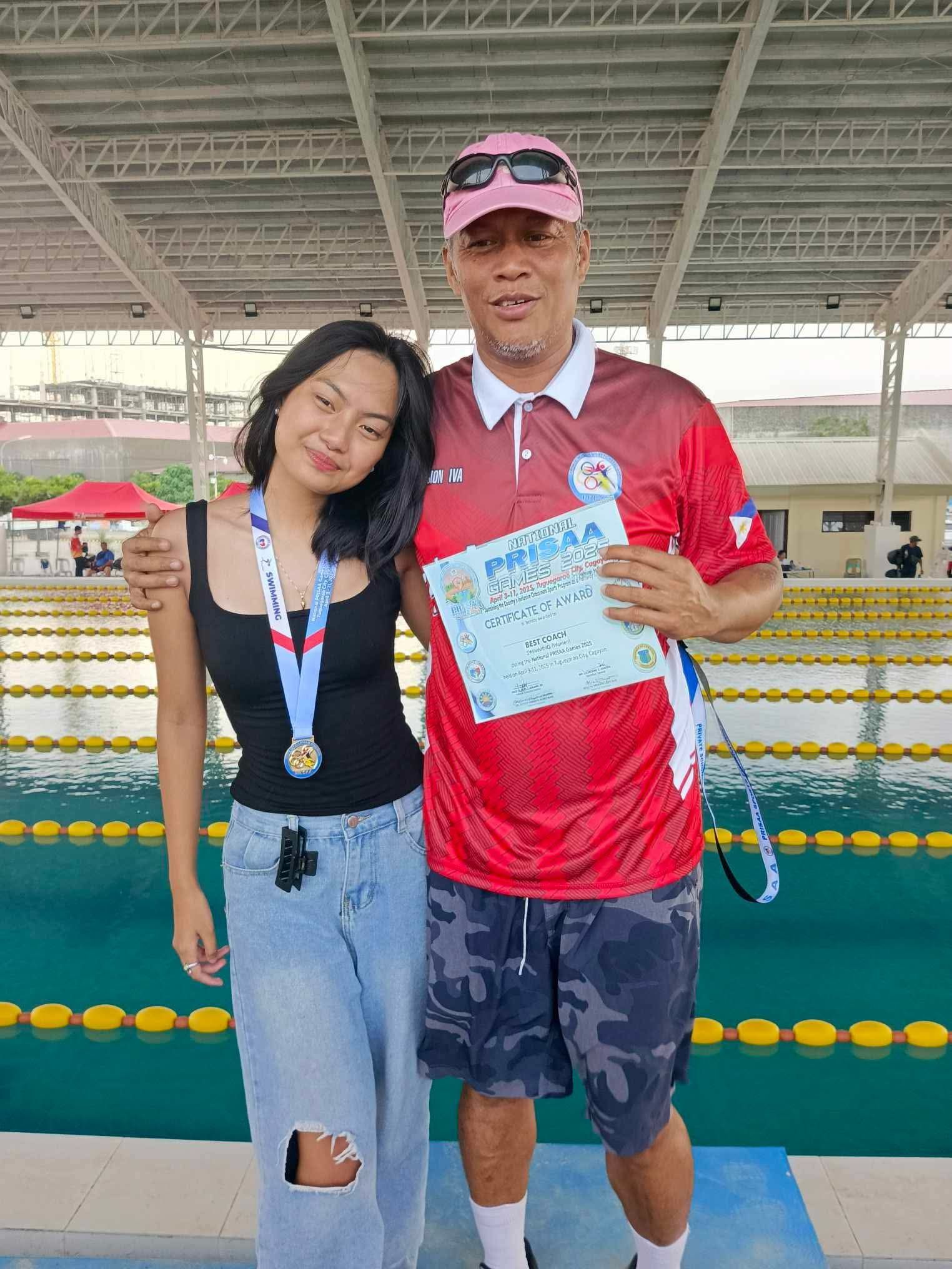 Two people posing by an indoor swimming pool, holding a certificate and medals.
