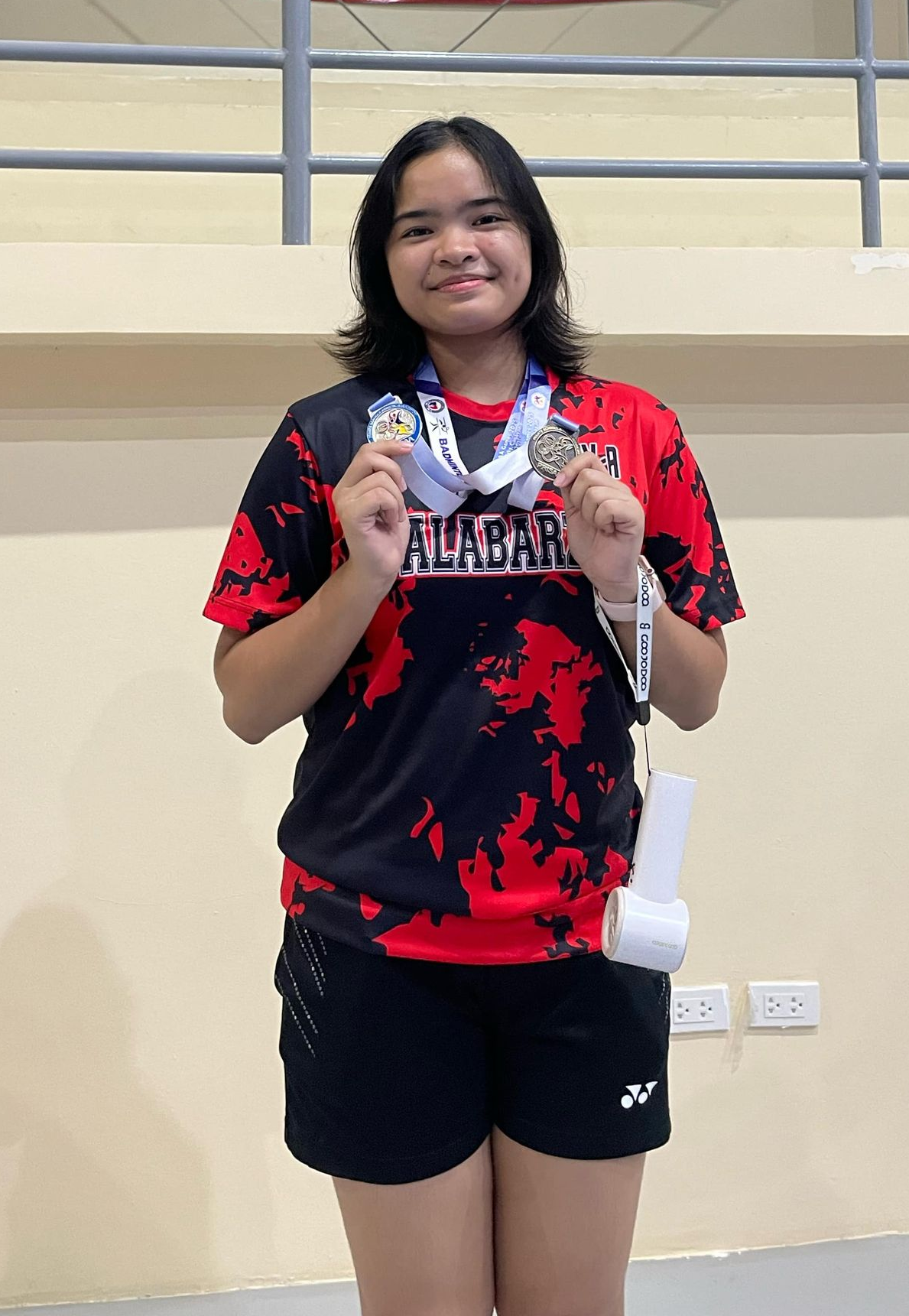 Person in a red-and-black jersey holding a medal, standing indoors and smiling at the camera.