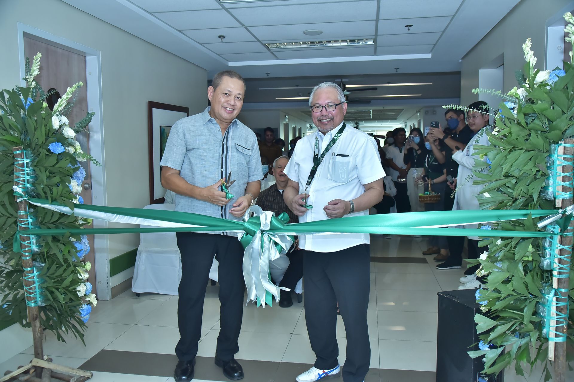 Two men cut a green ribbon at an indoor opening ceremony, with people watching behind them.