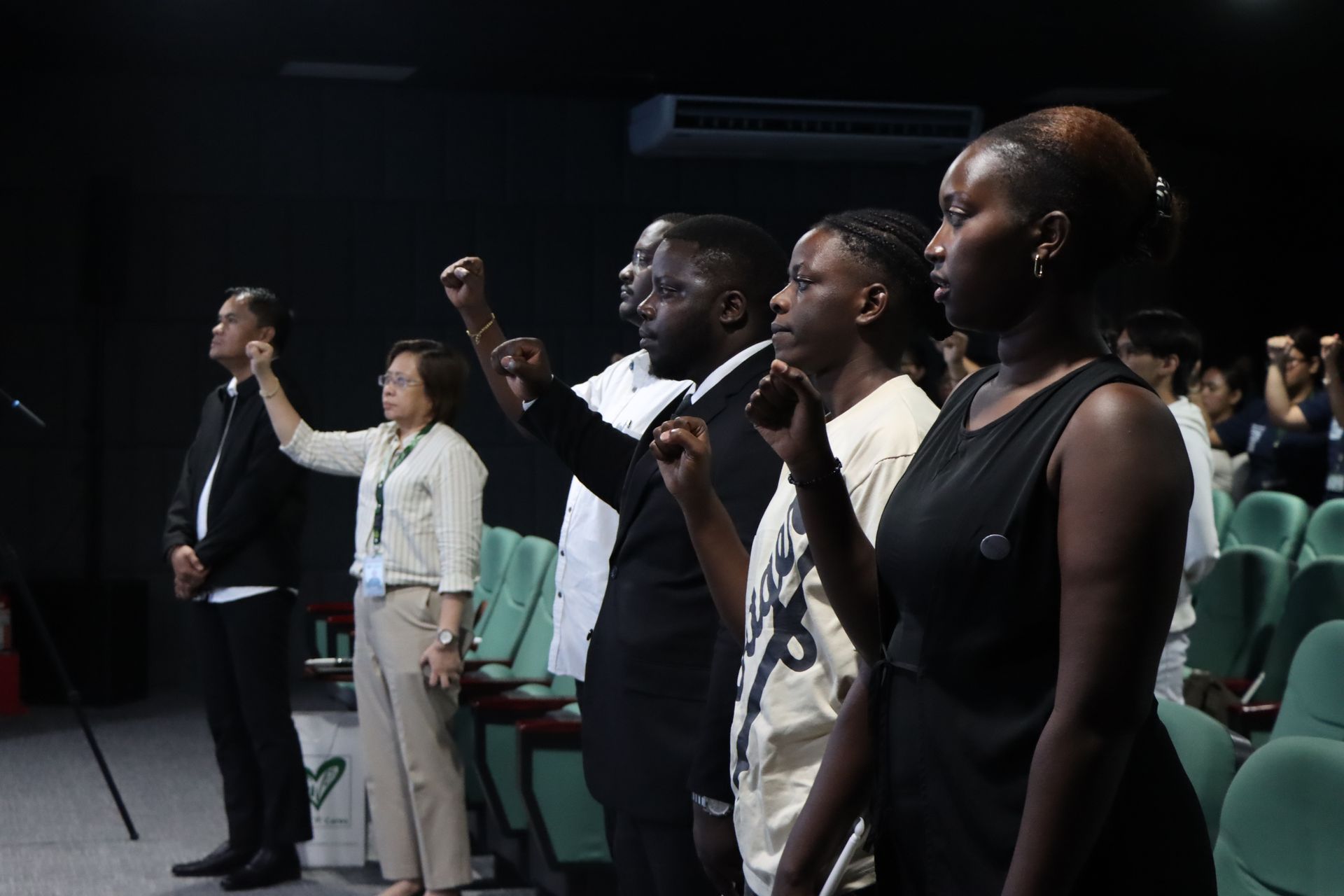Audience members stand and raise fists during a gathering in an auditorium.