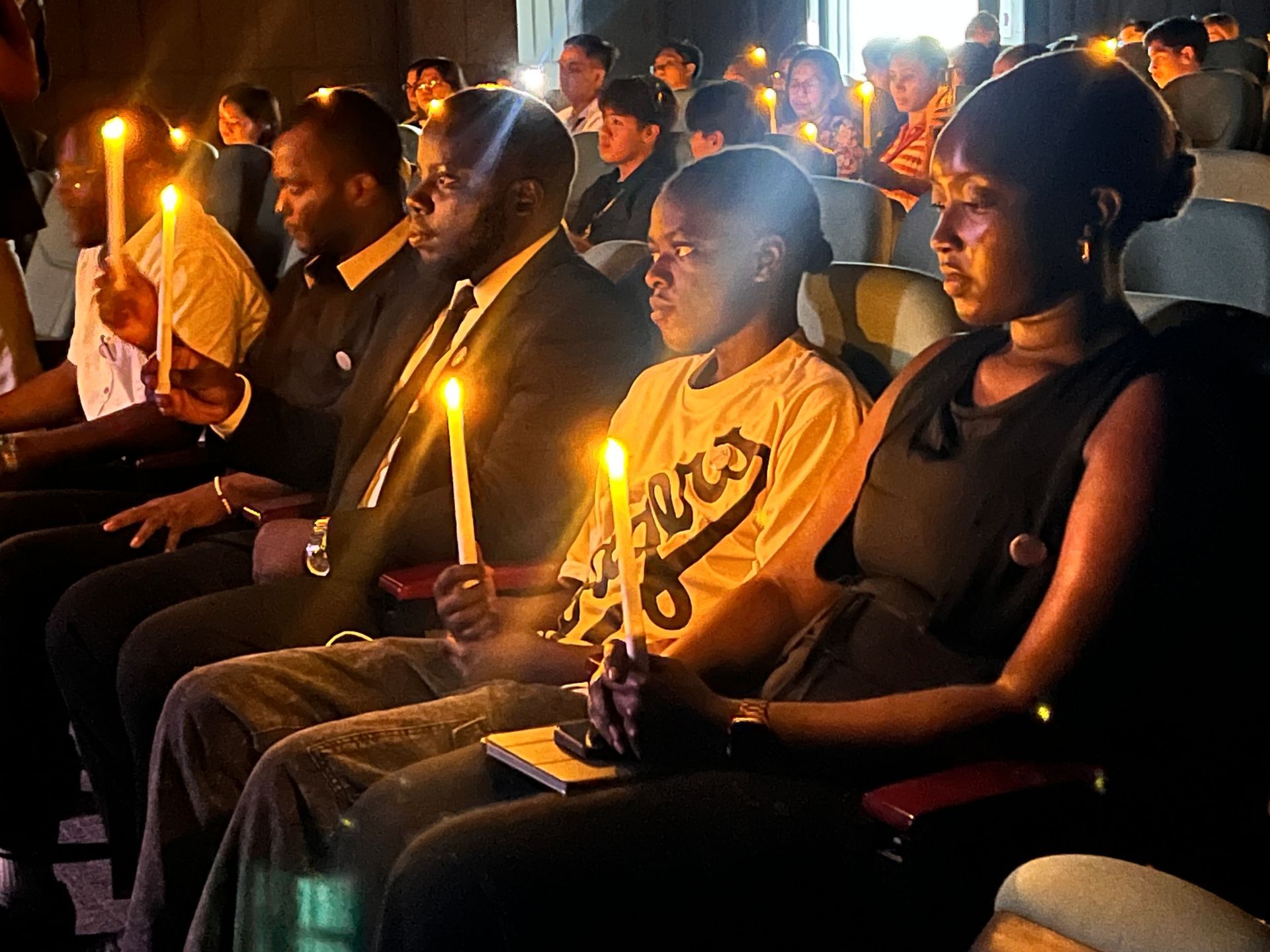 People seated in a candlelight vigil, holding lit candles in a dimly lit hall
