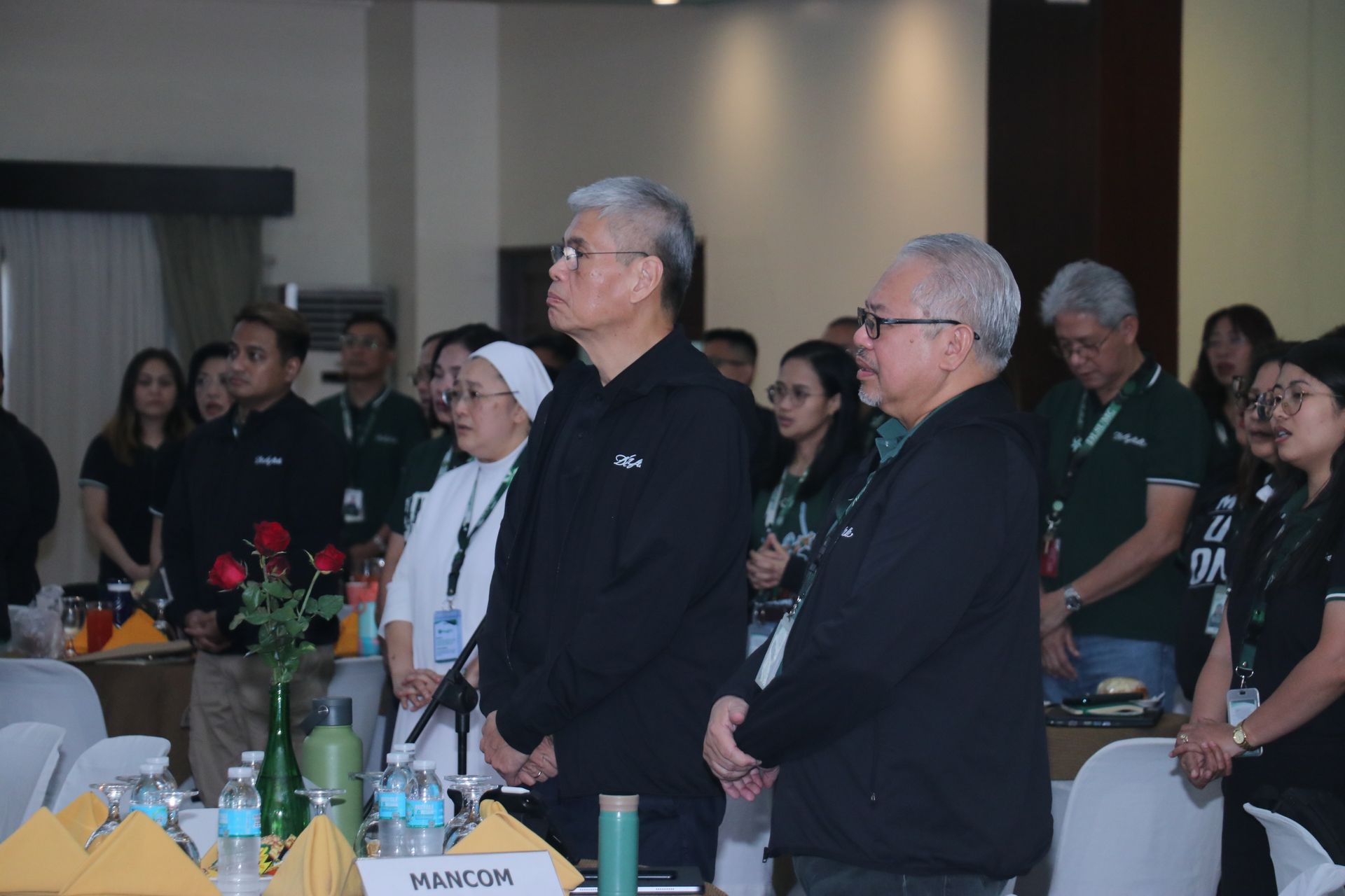 Attendees standing at tables during a formal indoor event, listening attentively.