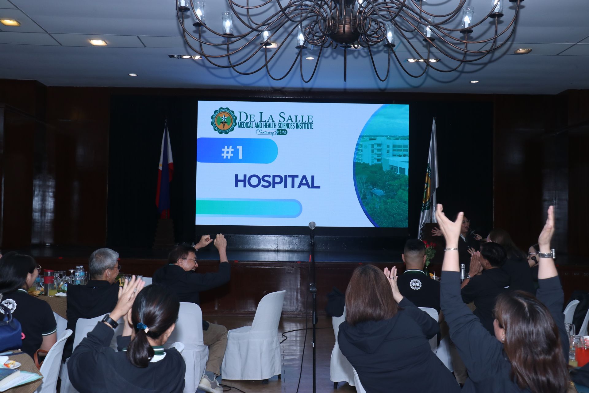 Conference audience facing a large screen that says “HOSPITAL” with a #1 graphic.