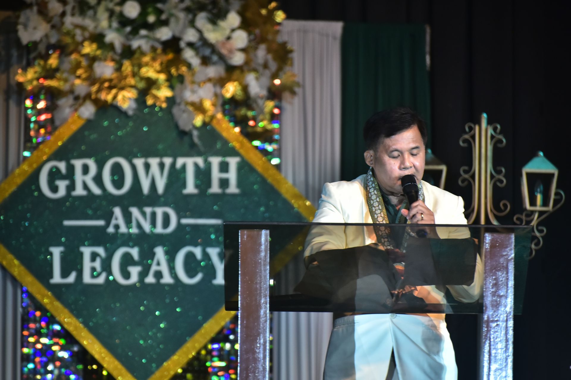 Speaker at podium during a “Growth and Legacy” event, with floral decor and lights in the background