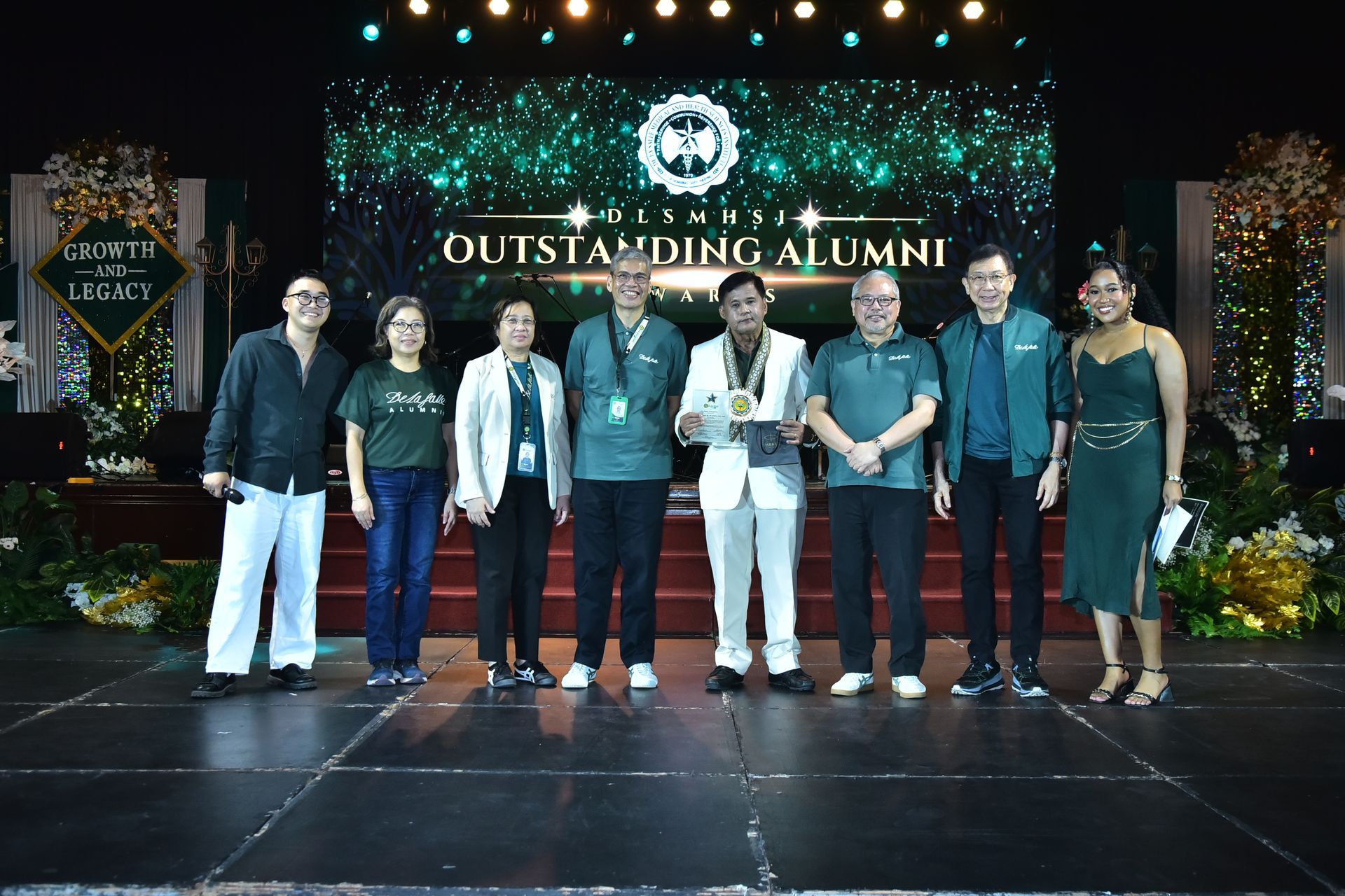Group of eight people on stage holding awards under an “Outstanding Alumni” sign.