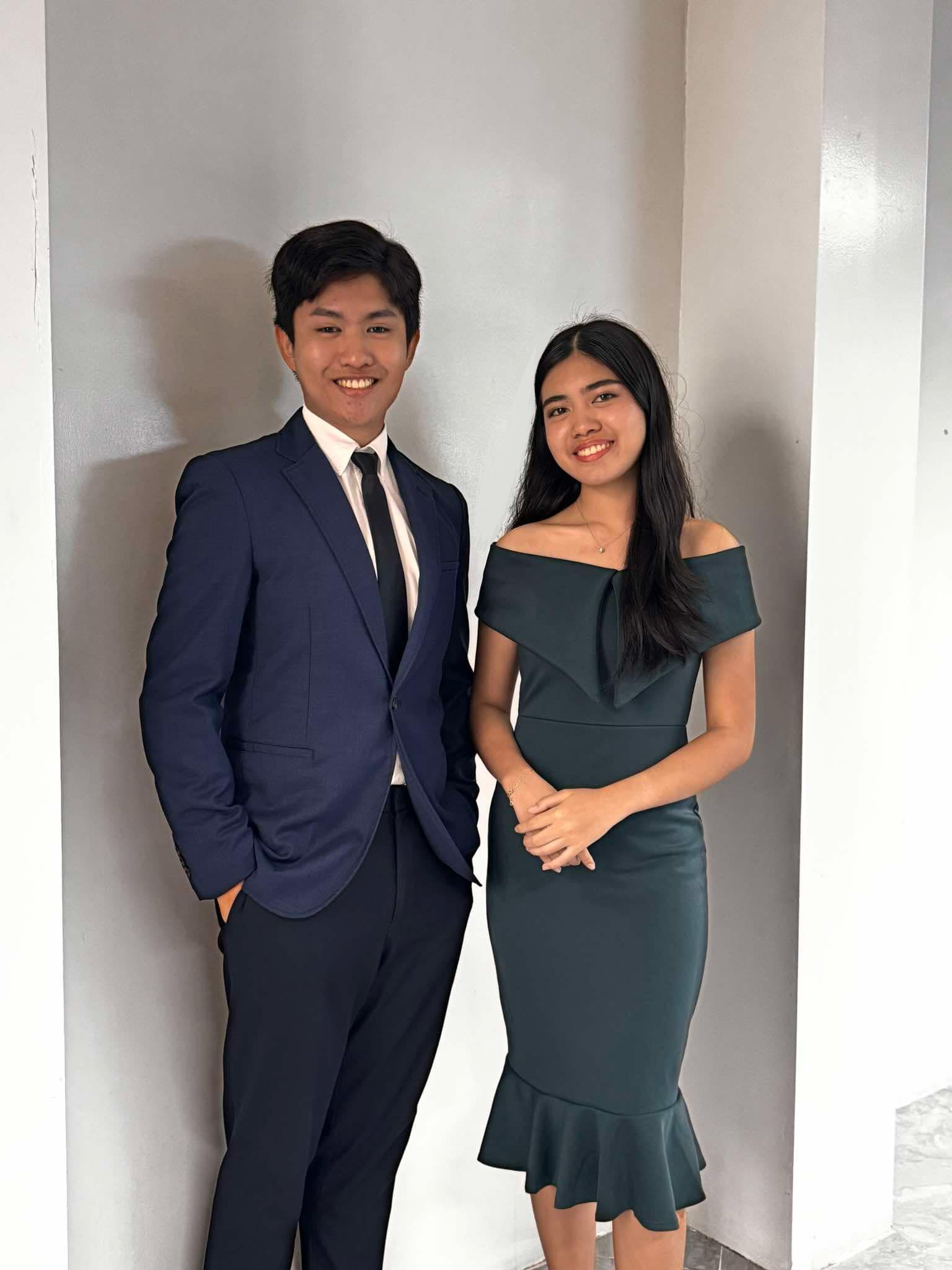 Two people in formal attire posing indoors against a plain light wall.