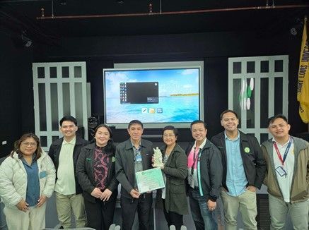 Group of eight people posing on a stage in front of a presentation screen and flags
