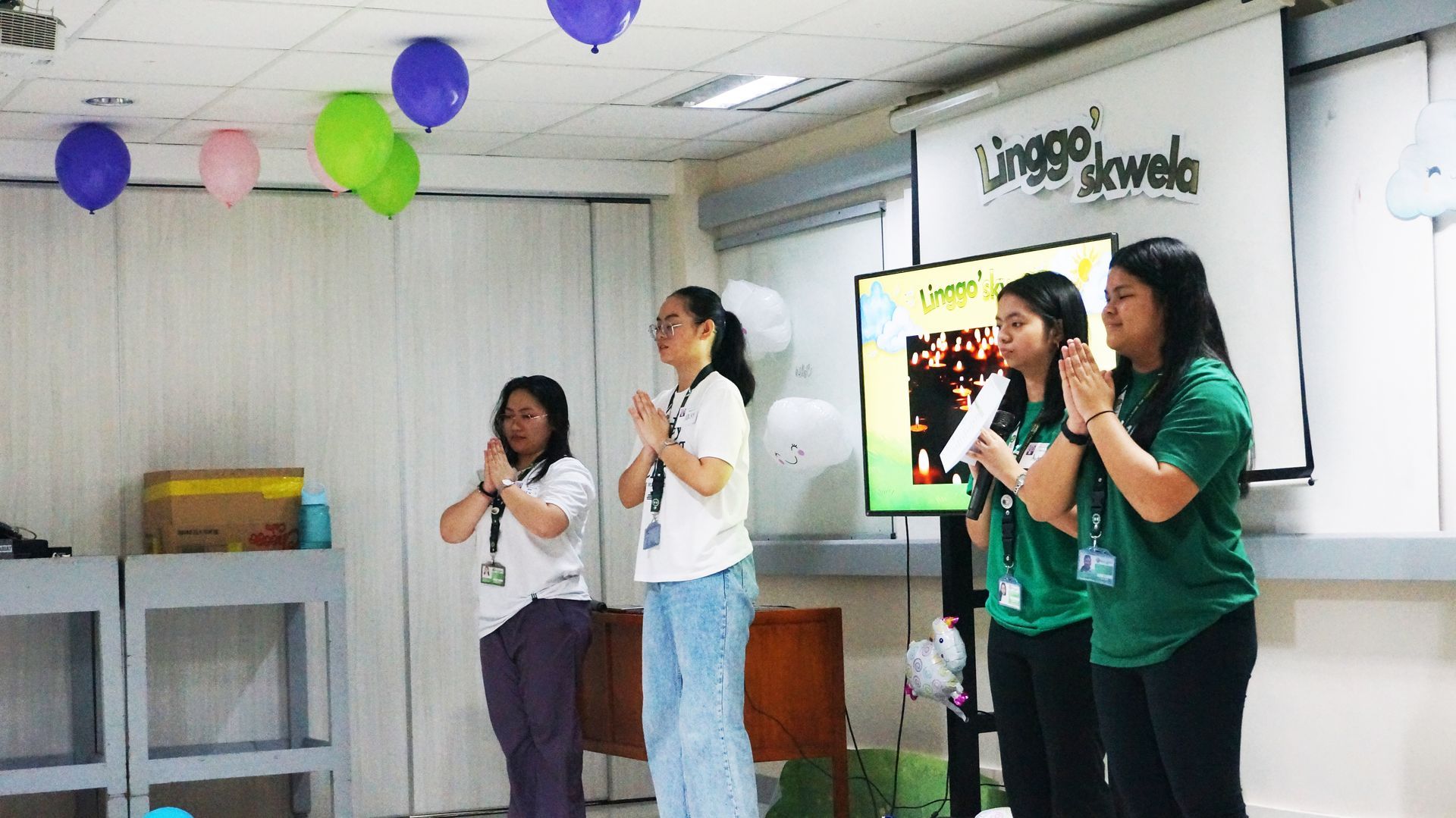 Four people stand on a stage singing into microphones in a bright room with colorful balloons and a TV screen.