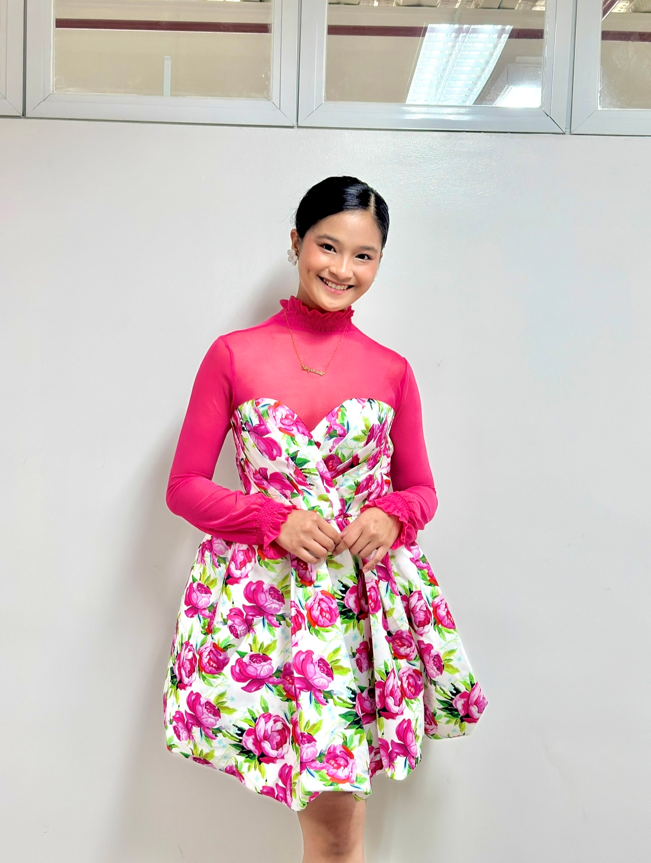 Person in a pink-and-floral dress smiling and standing indoors against a plain wall.
