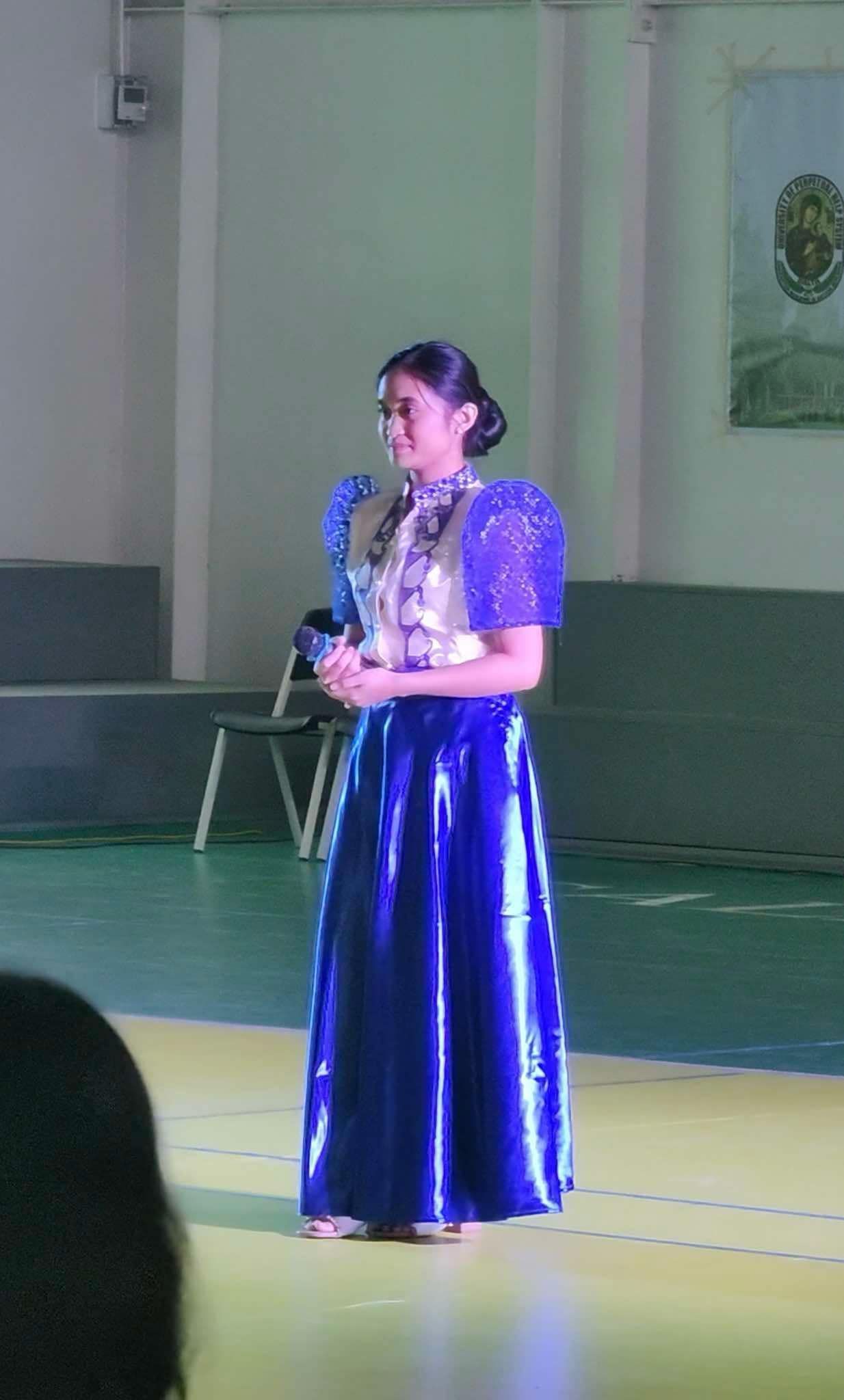 Woman in a shiny blue dress standing on a stage holding a microphone and looking left