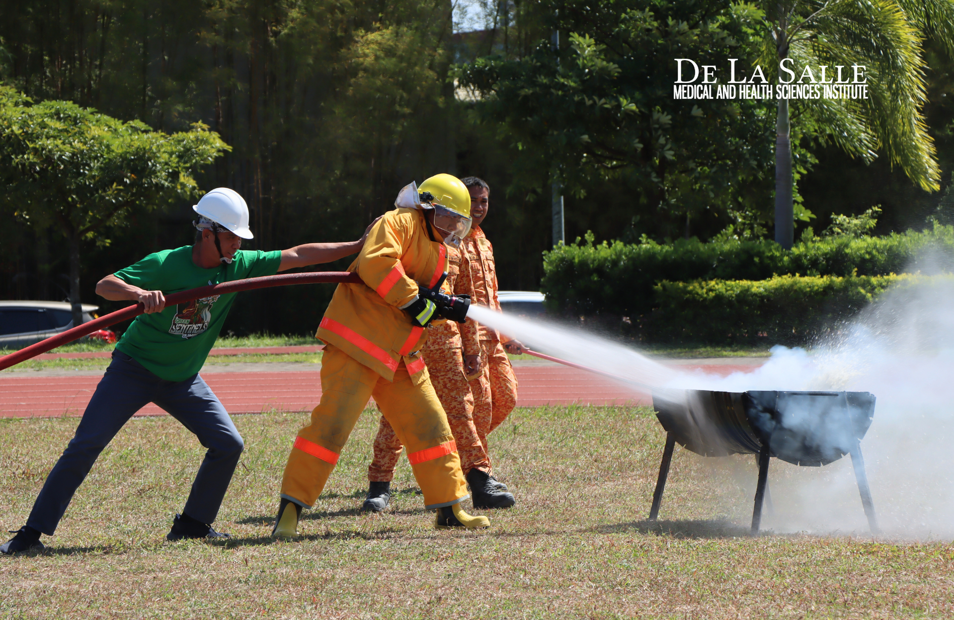 Three firefighters in helmets spray water on a smoking black chair outdoors during a fire drill
